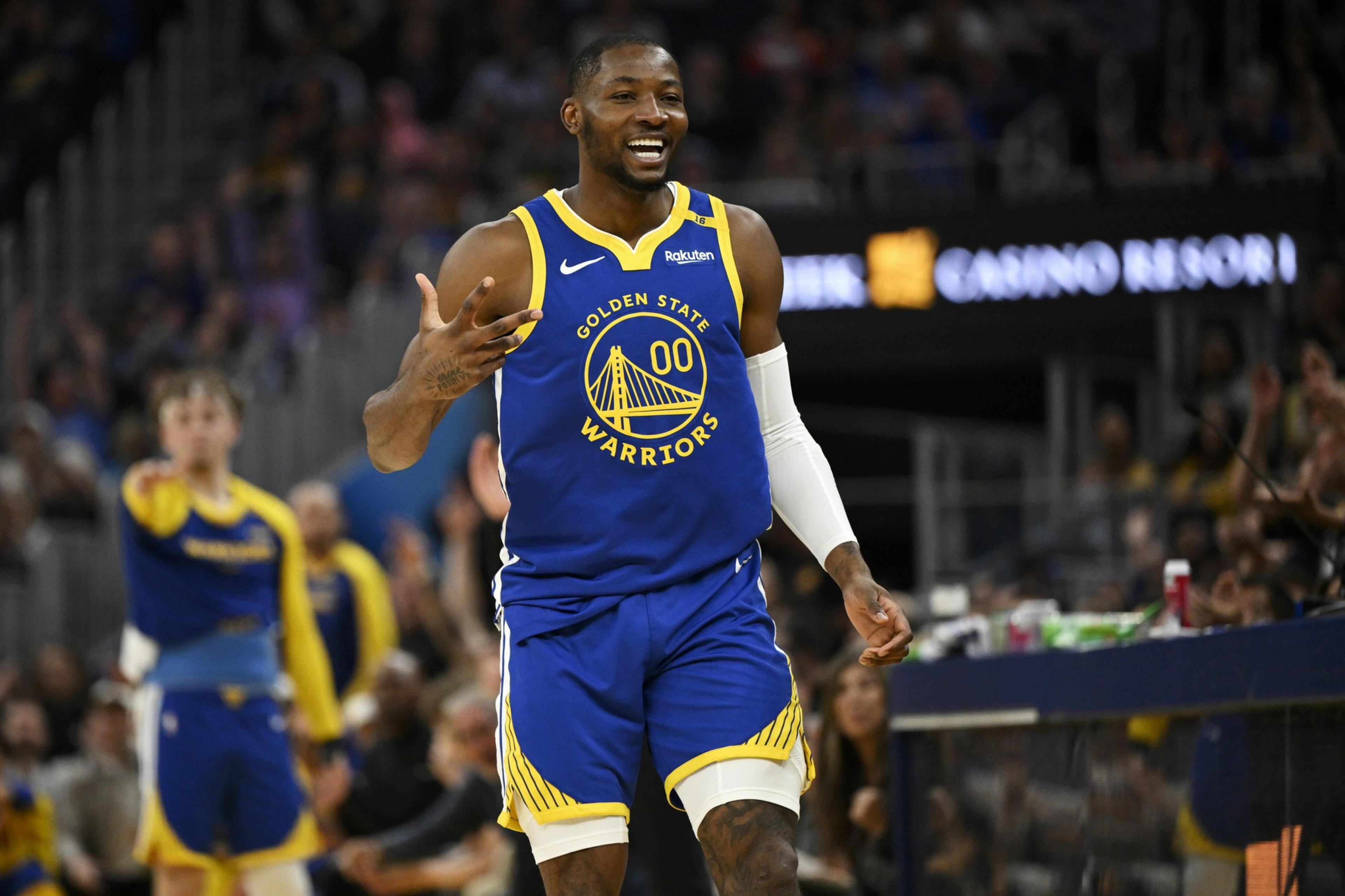 A basketball player in a Golden State Warriors jersey is on the court, smiling and gesturing. The background shows a blurred crowd and part of the court.