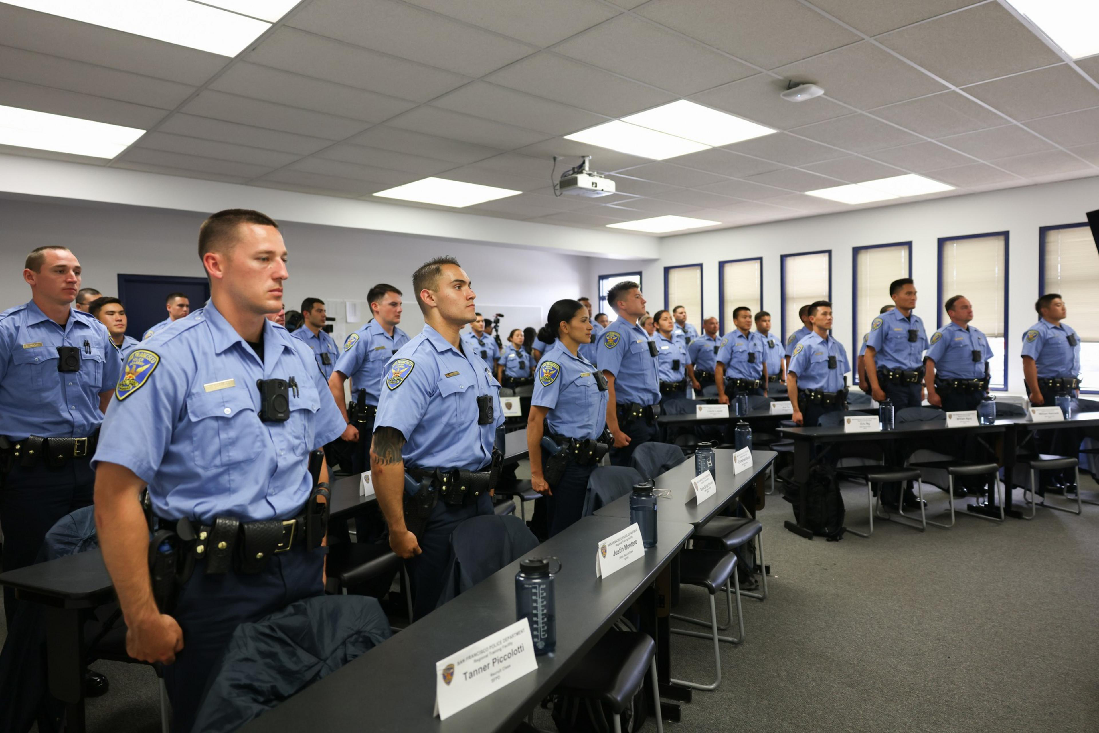 A group of uniformed officers stand attentively in a classroom setting. They wear light blue shirts and dark pants, with name tags visible on the desks in front.