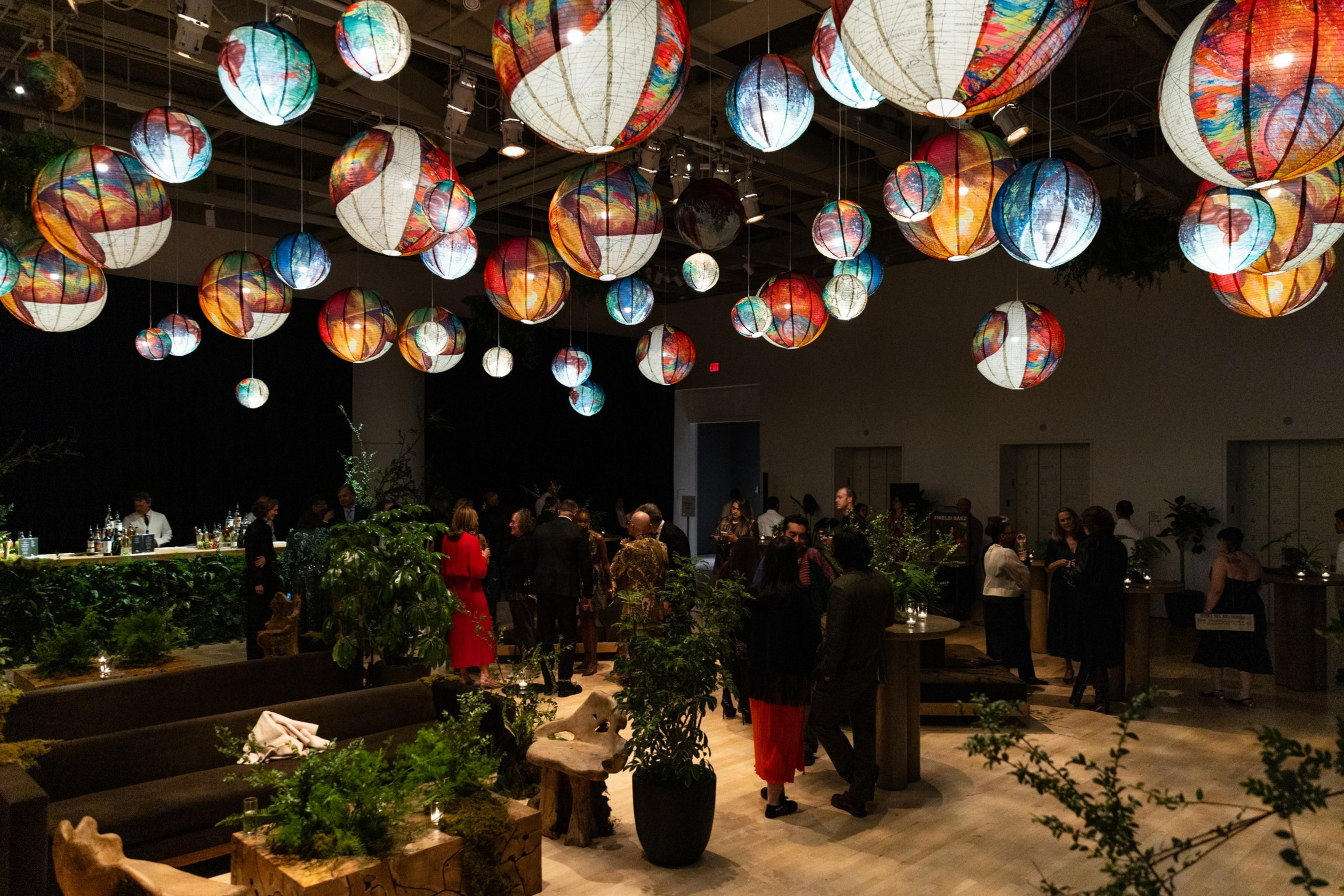 The image shows a lively indoor event with colorful, spherical lanterns hanging from the ceiling. Guests are mingling amidst lush greenery and artful seating.