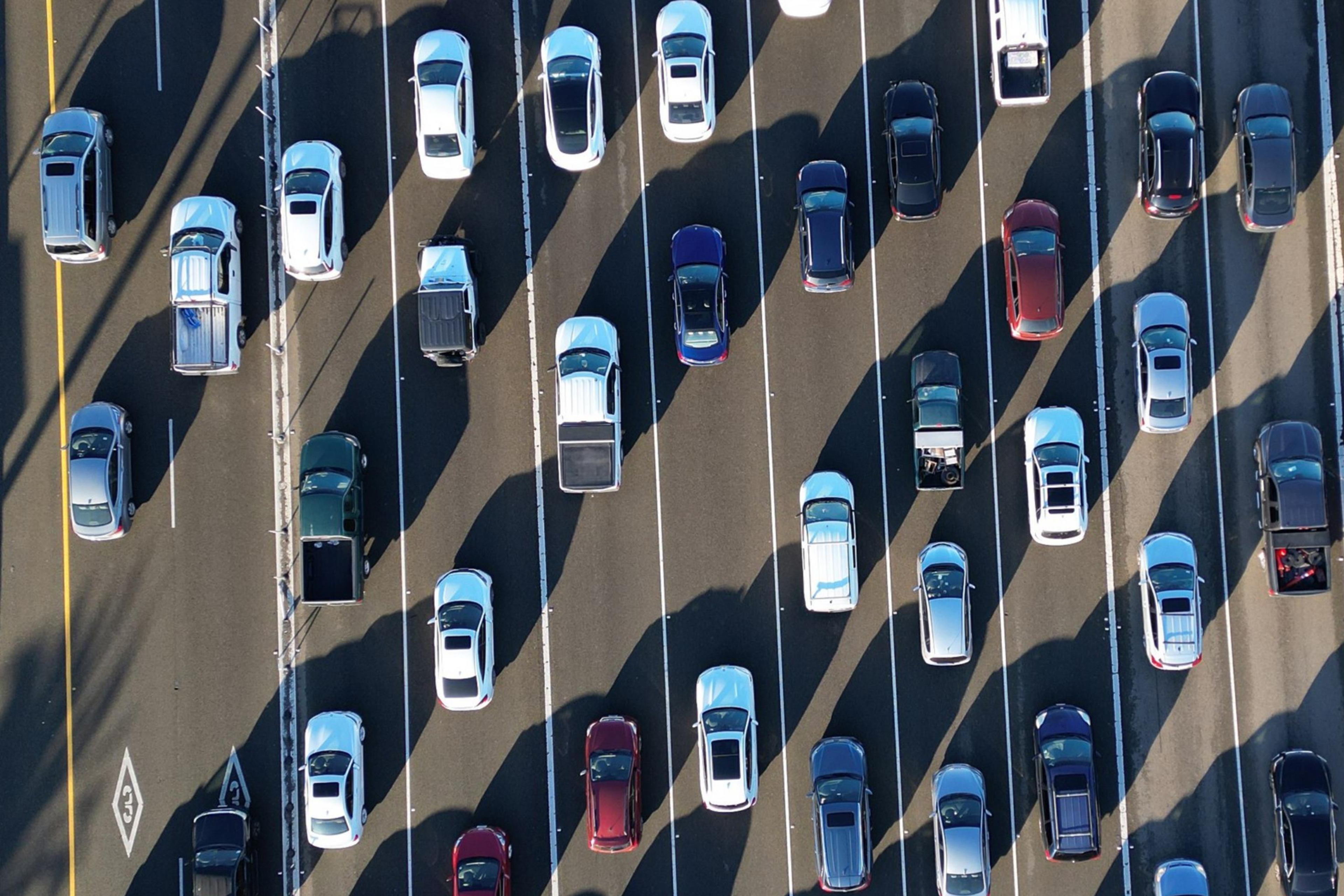 The image shows multiple lanes of traffic from above, with rows of cars and trucks in various colors, creating a pattern of parallel lines and shadows.
