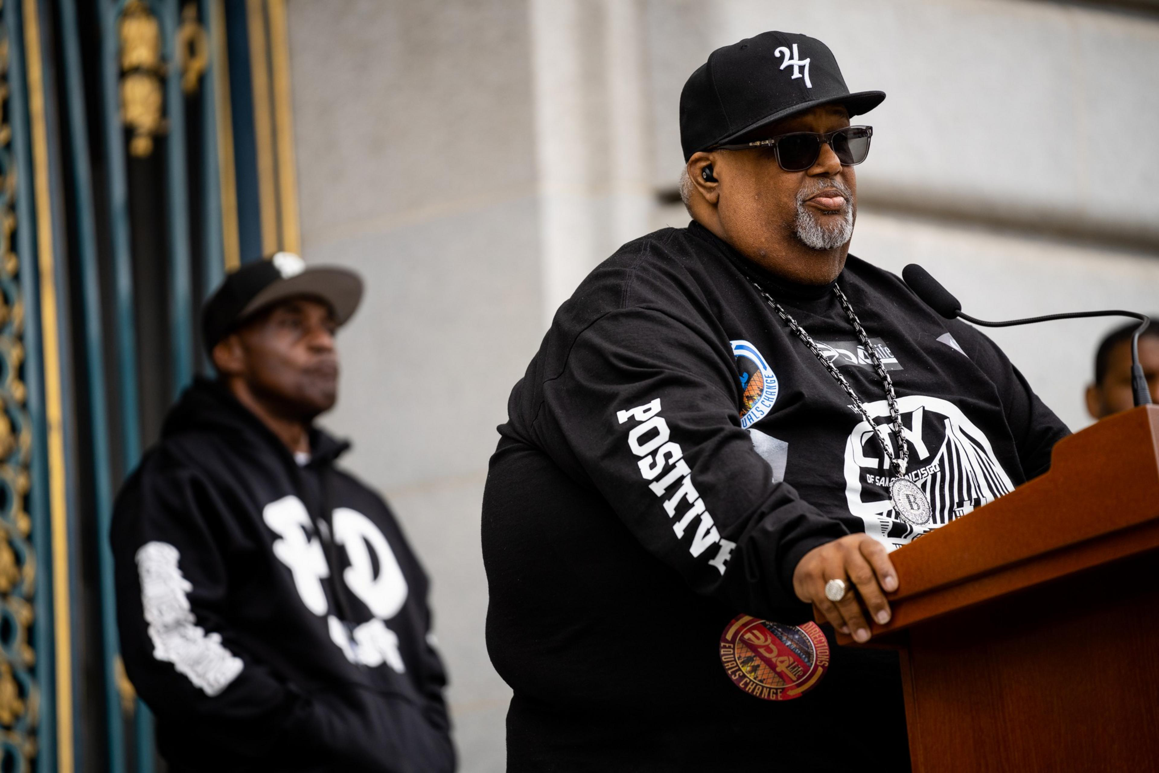 A man in sunglasses and a baseball cap speaks at a podium with a serious expression; another man stands in the background wearing a hoodie and cap.