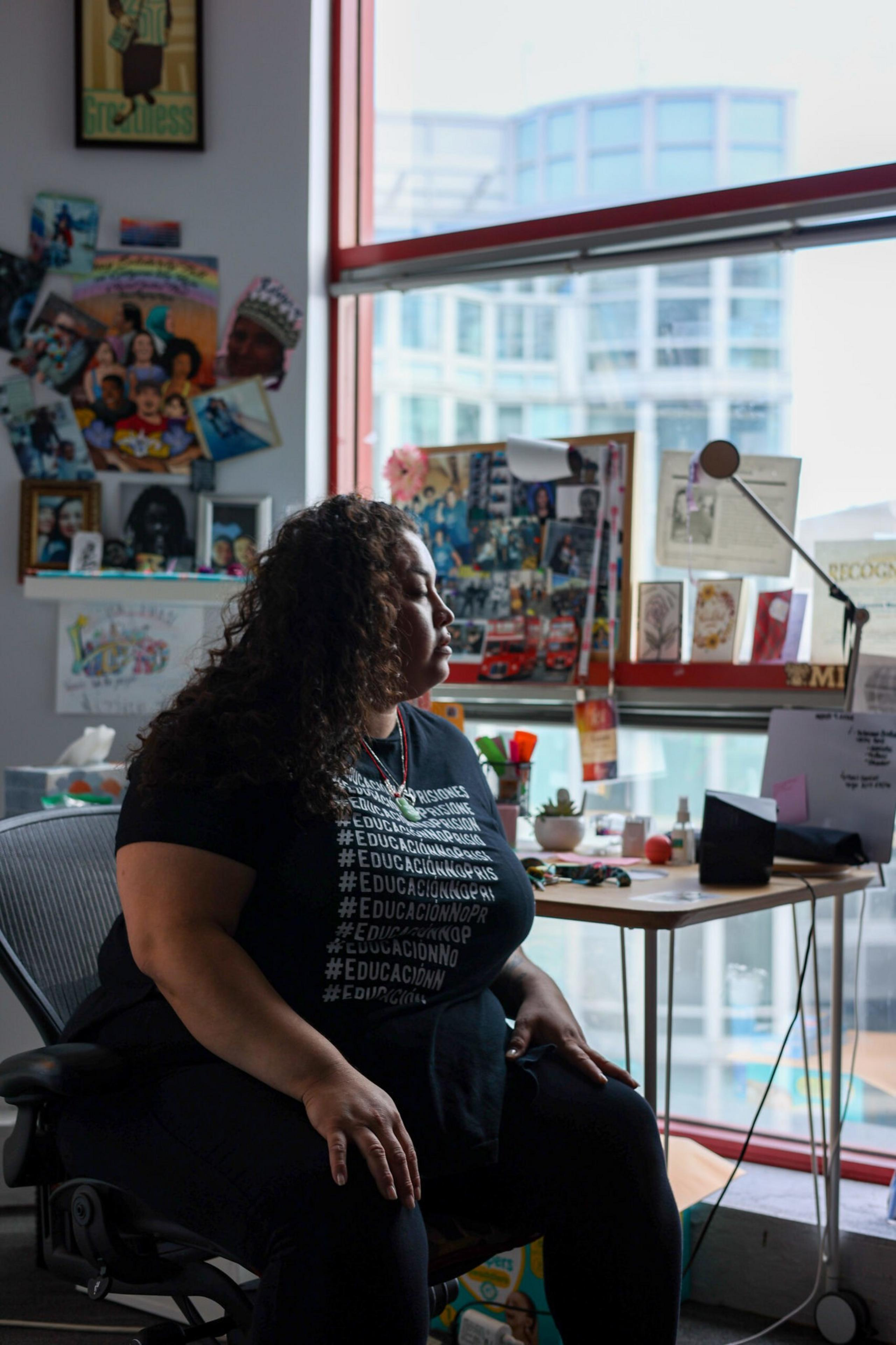 A woman with curly hair sits in an office chair, looking thoughtfully out a window beside a desk cluttered with papers and personal items.