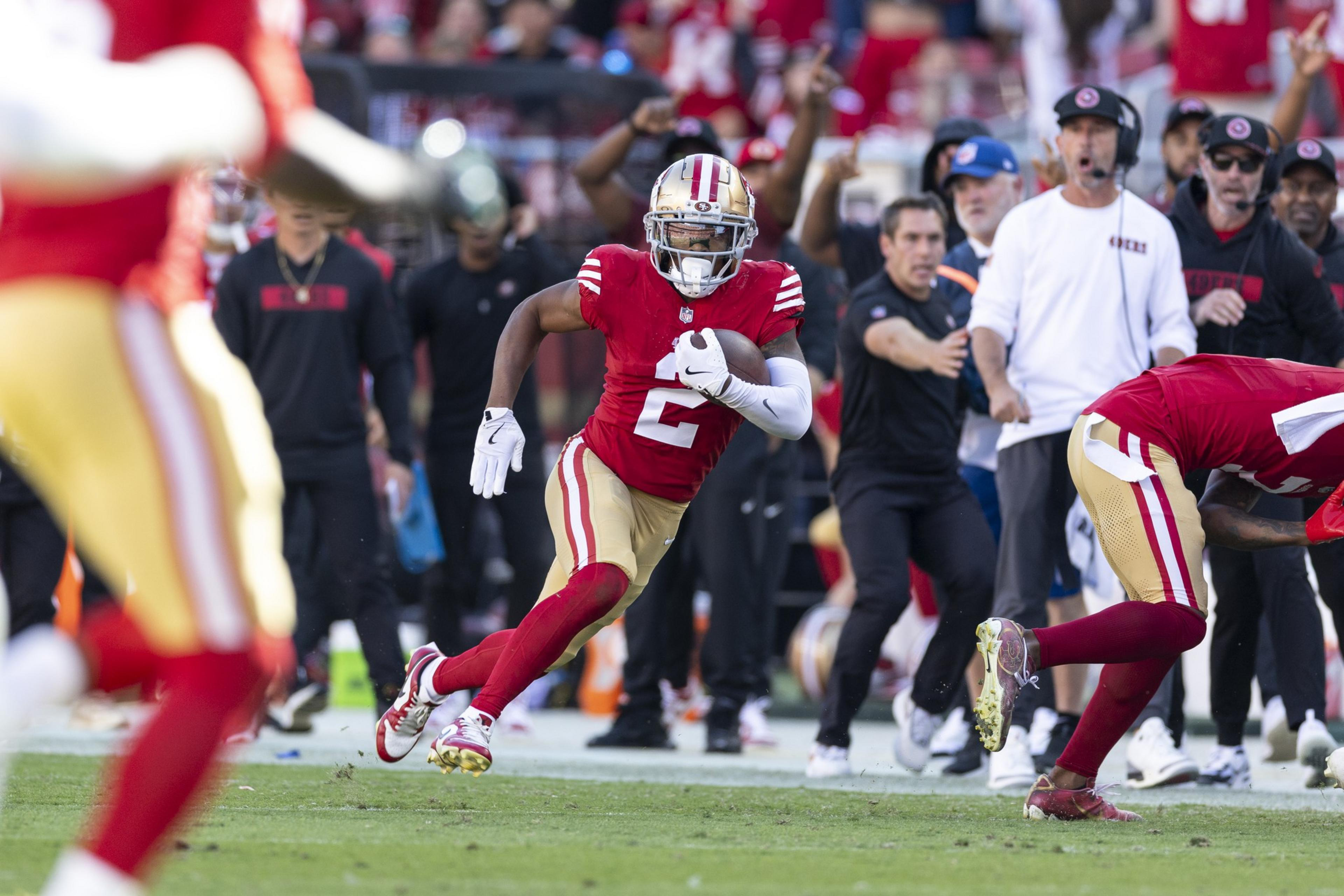 A football player in a red jersey runs with the ball on a field, closely followed by teammates. Coaches and staff on the sidelines react in the background.