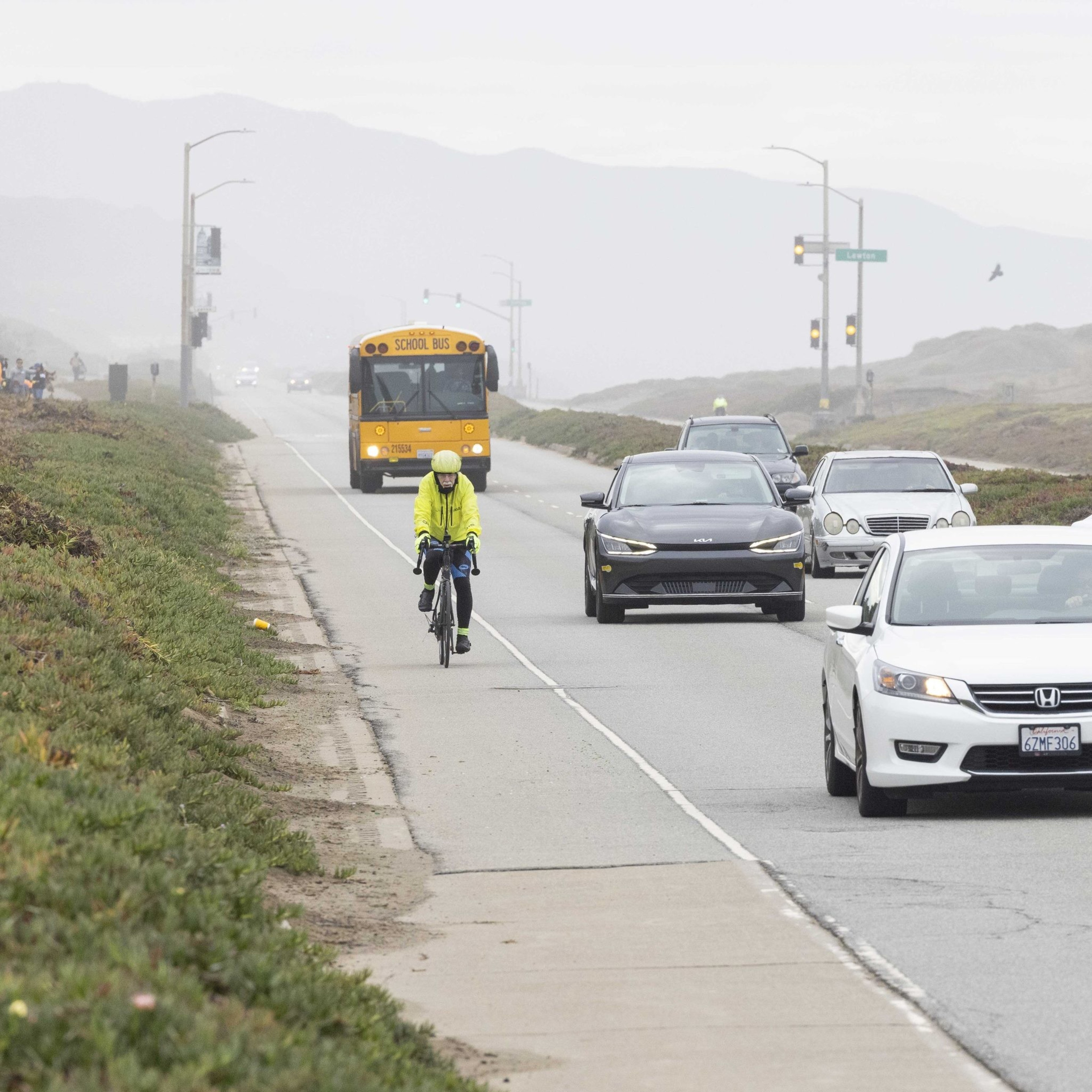 A cyclist in a yellow jacket rides along a roadside bike lane. Cars and a yellow school bus travel beside him on a foggy day.
