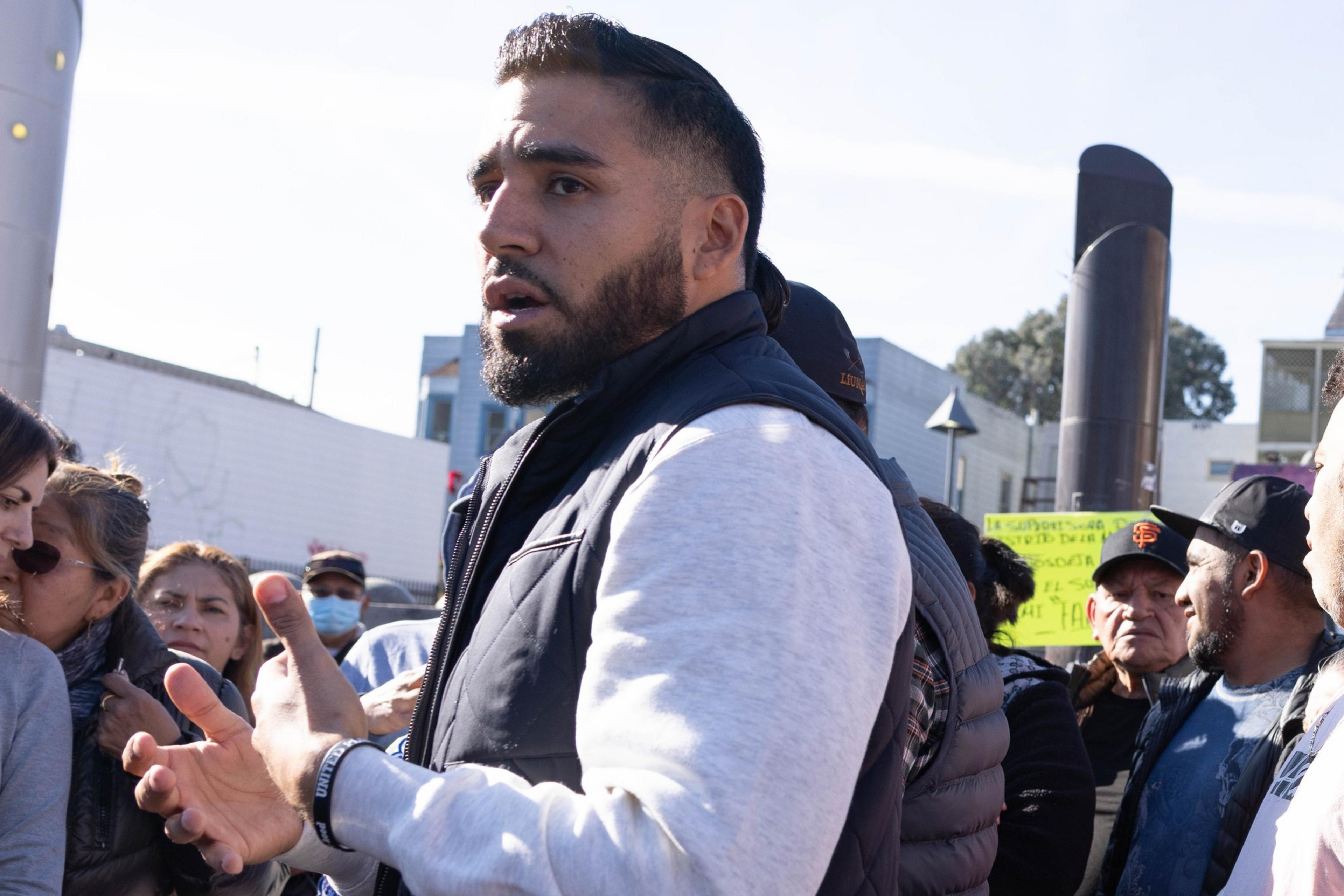 A man gestures while speaking to a group of people outdoors, possibly at a gathering or event.