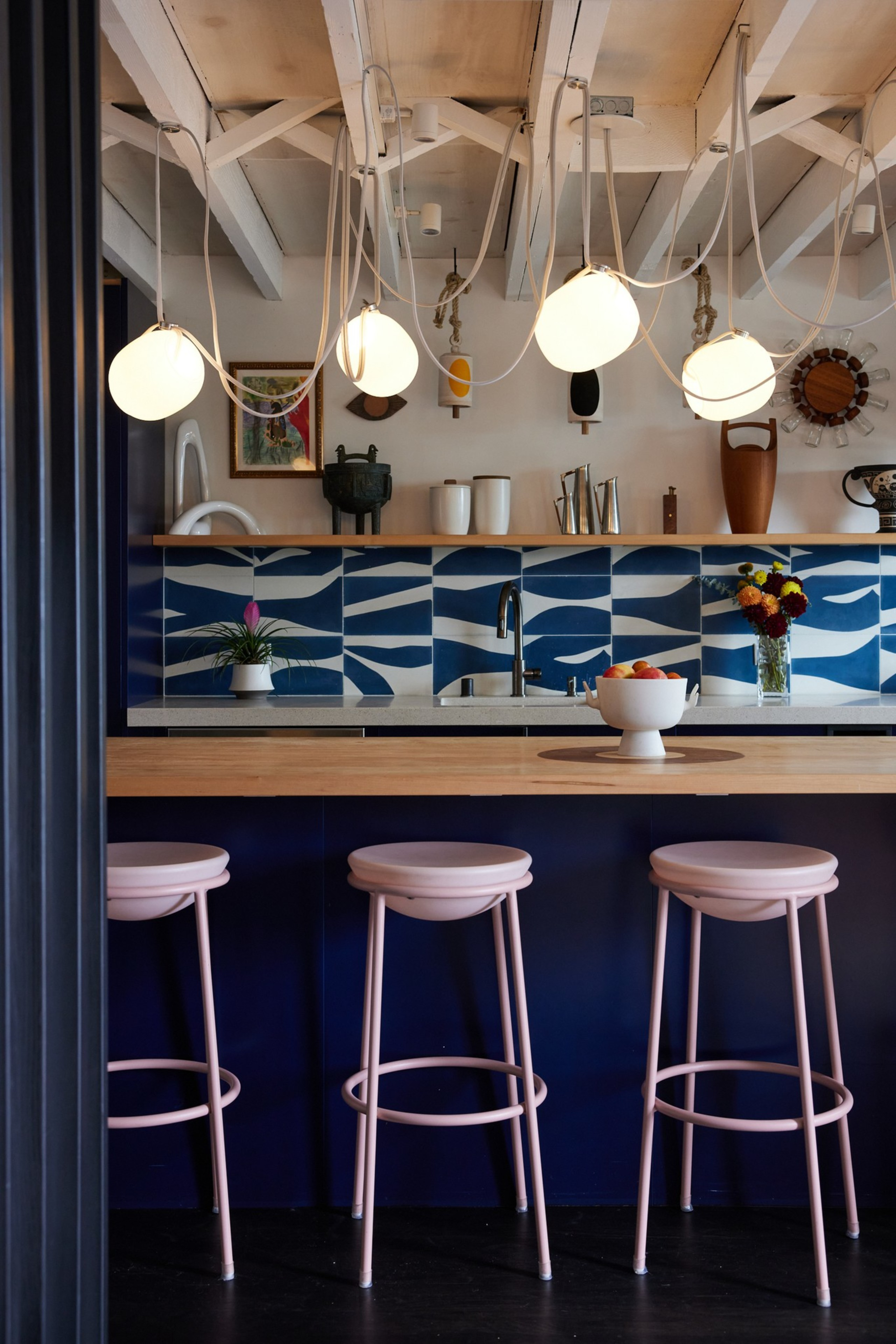 This image shows a stylish kitchen with modern light fixtures, blue patterned backsplash tiles, a wooden countertop, pink stools, and various decor items.
