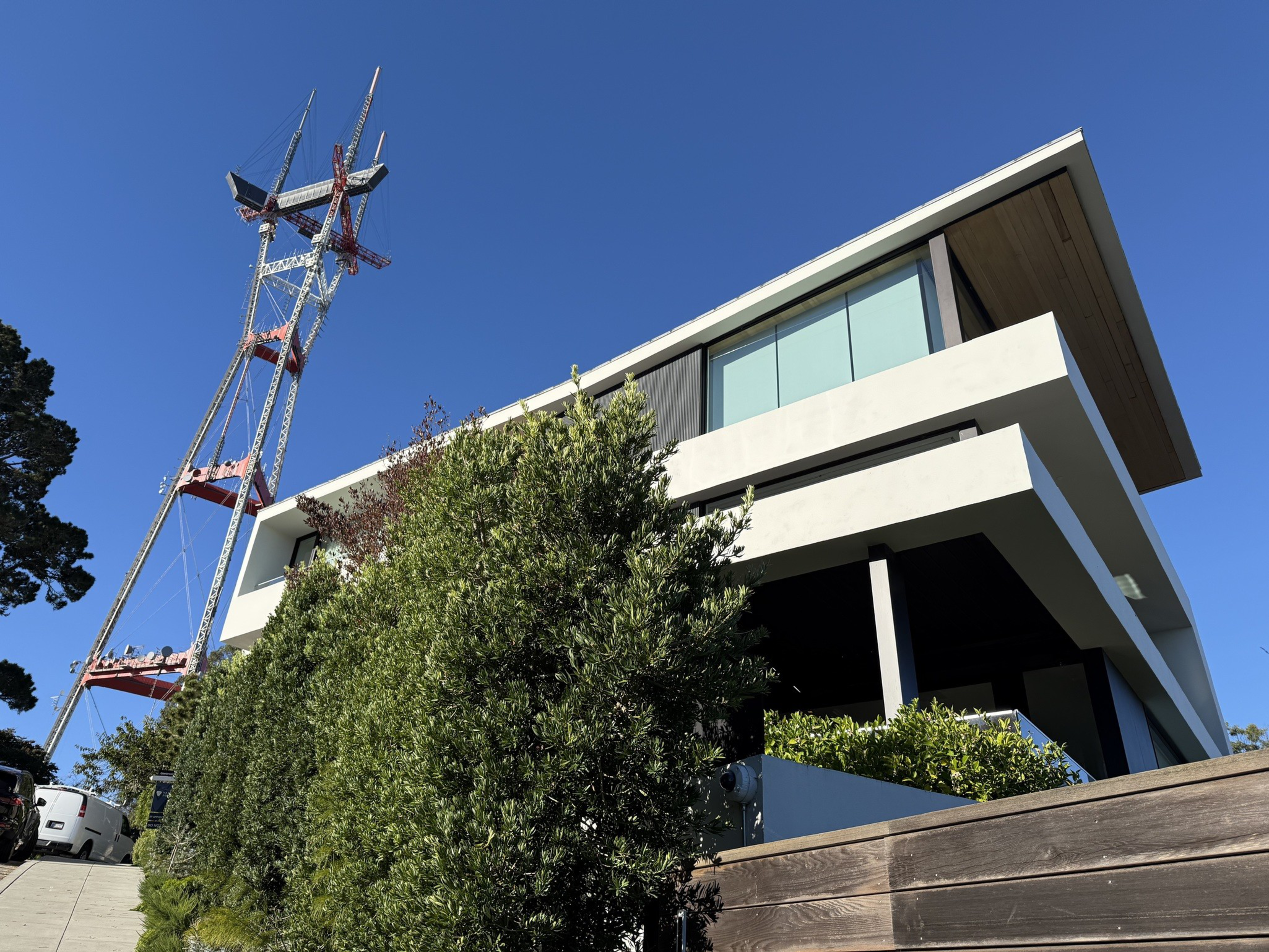 A modern, white and black two-story building stands beside a tall red and white radio tower, surrounded by dense green shrubs under a clear blue sky.