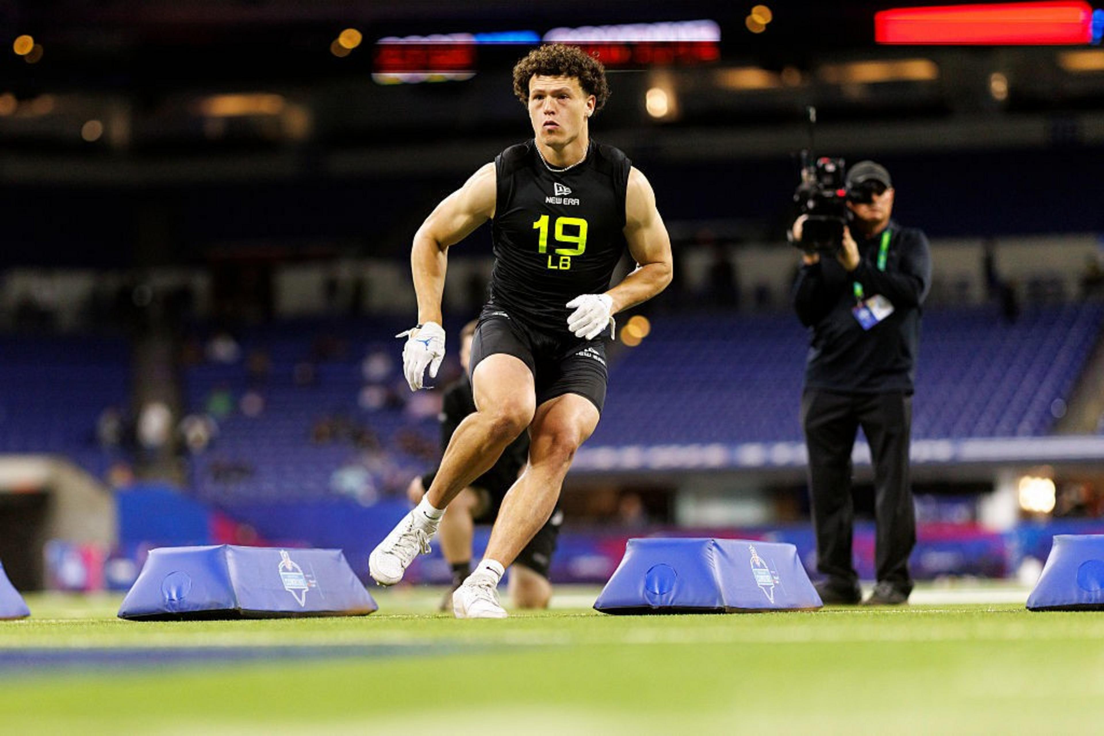 A football player in a black outfit and gloves is running between blue cones on a field indoors. A person with a camera is filming the action.