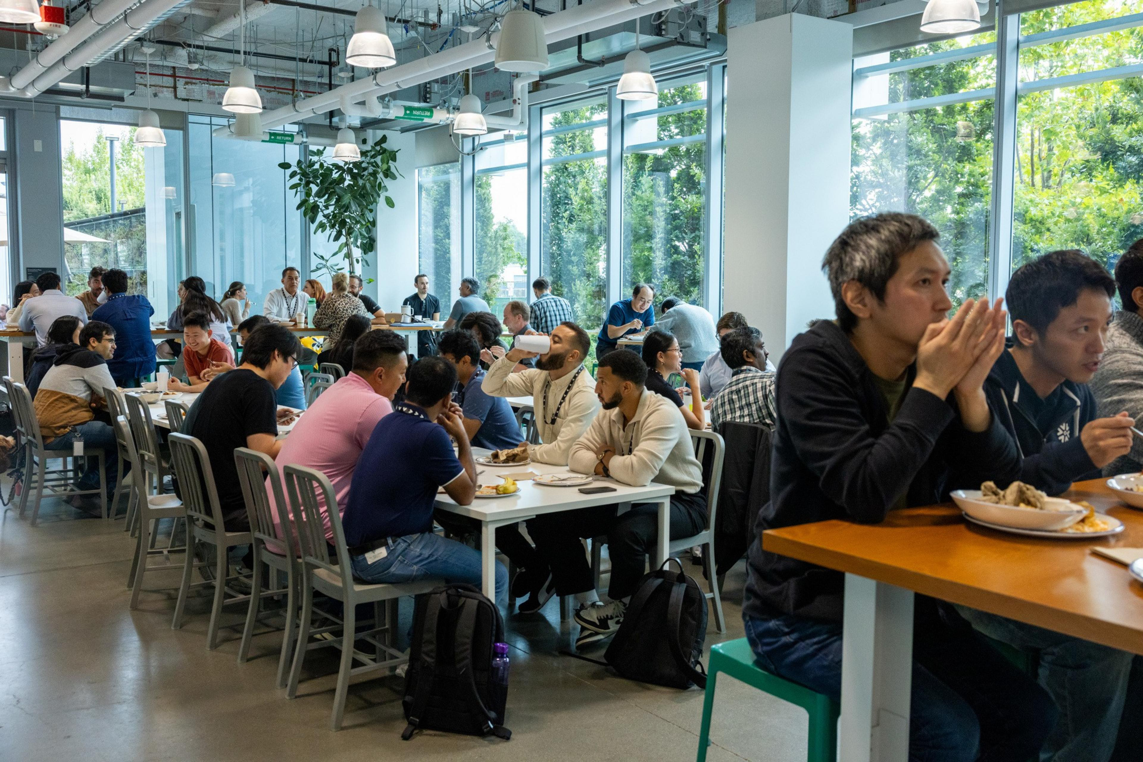 People sit closely at tables in a bright cafeteria with large windows, eating and talking in a busy, modern indoor space.