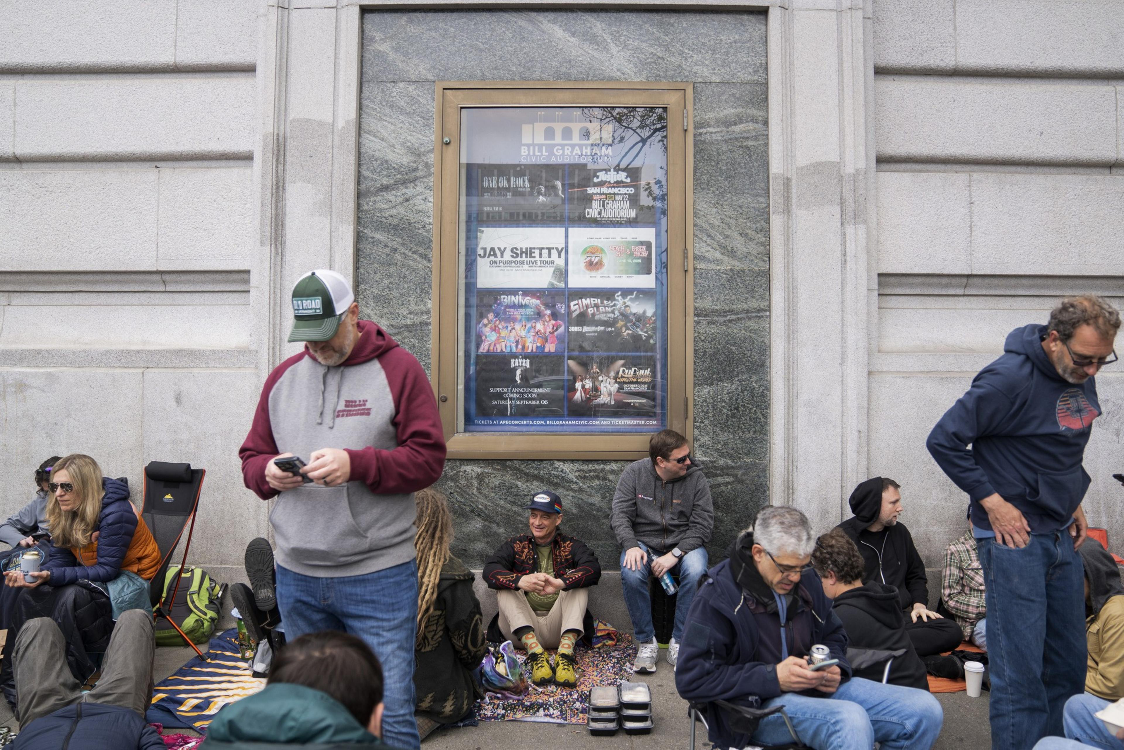 A group of people sits or stands outside a building with a poster in a frame. Some use phones and others chat, creating a casual, social atmosphere.