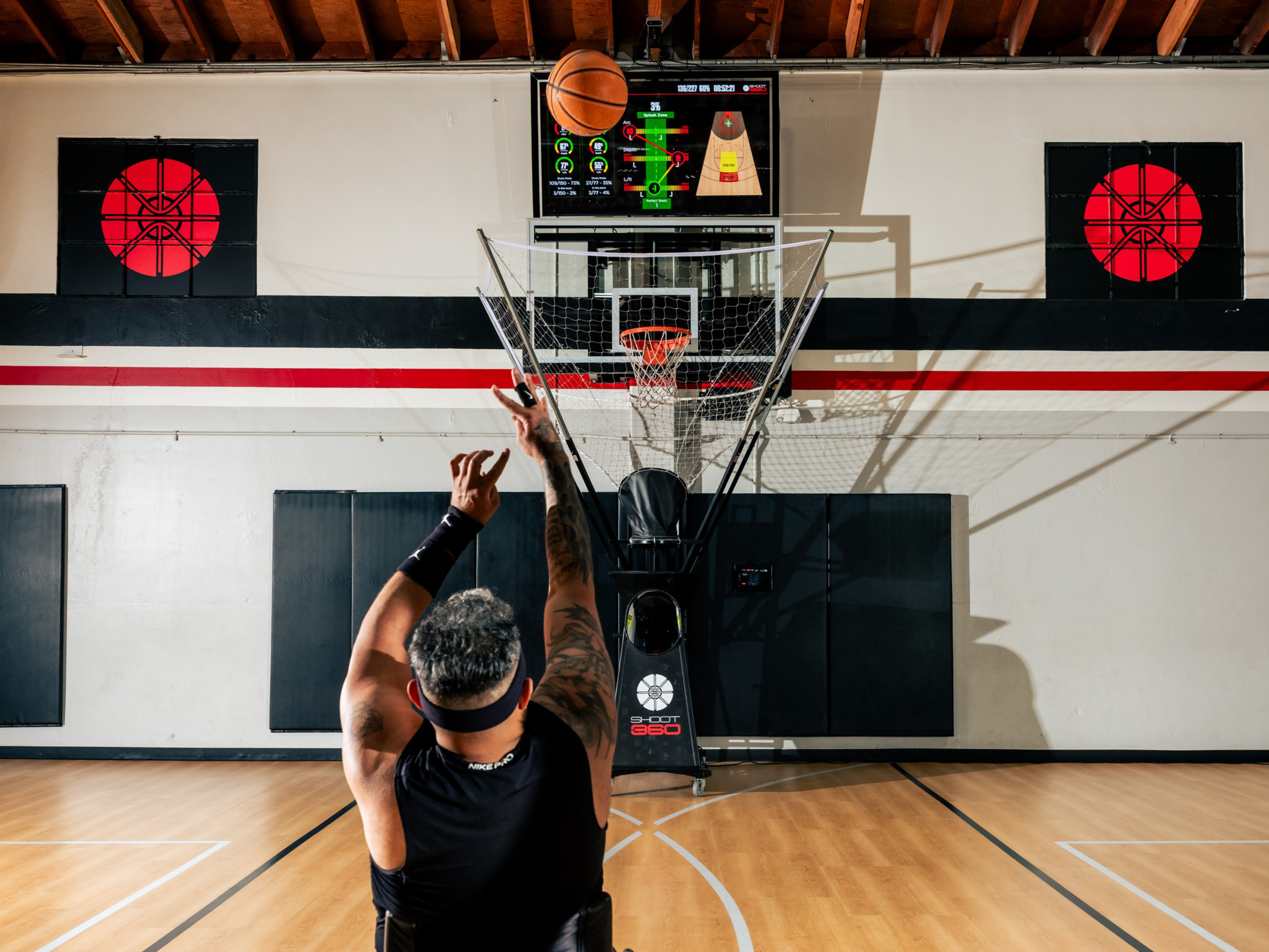 A man with a tattooed arm shoots a basketball toward a hoop equipped with a shot-tracking net and a digital scoreboard above it in a gym.