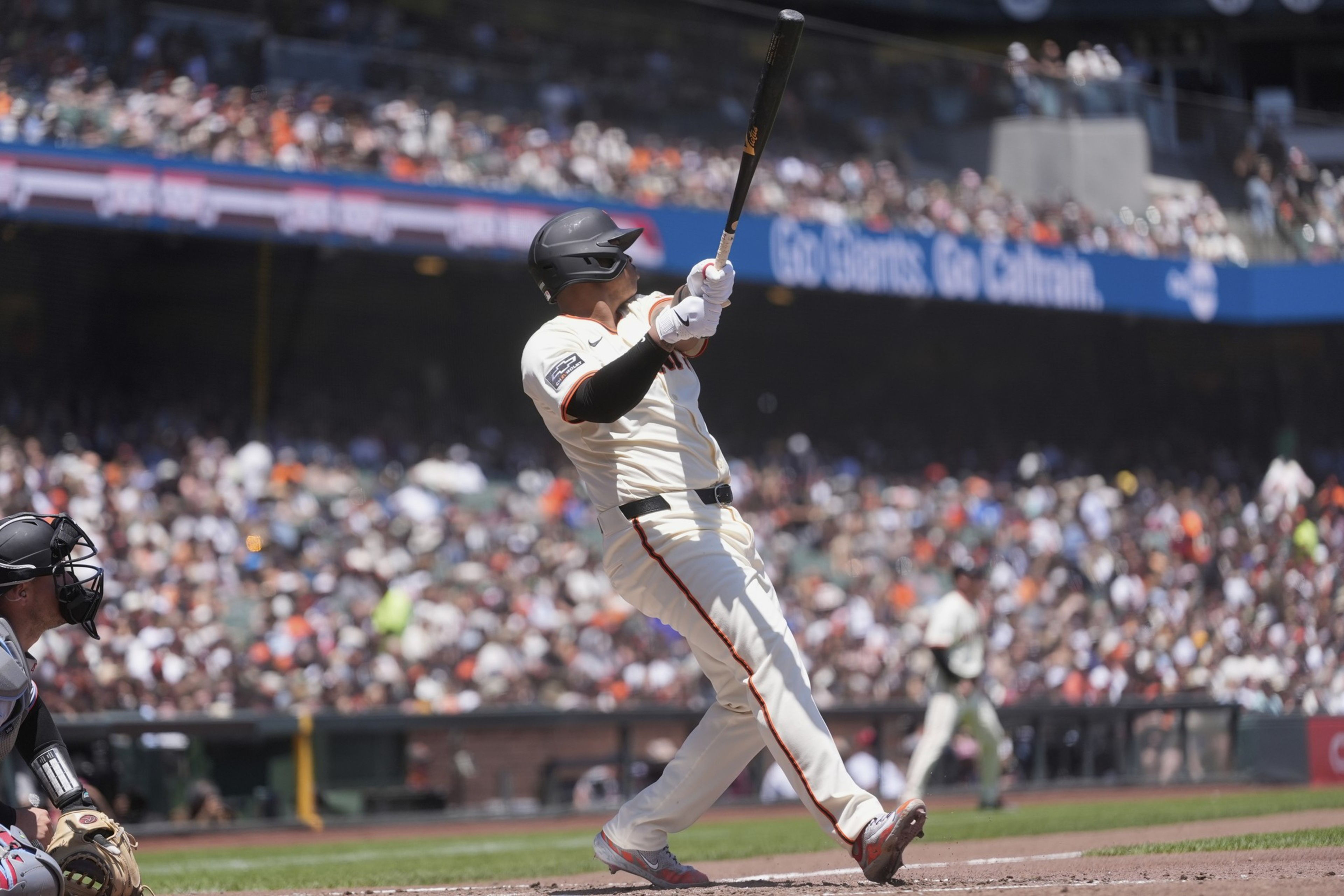 A baseball player, wearing a white uniform and black helmet, swings a bat at a stadium. A catcher crouches nearby and a large crowd fills the stands.