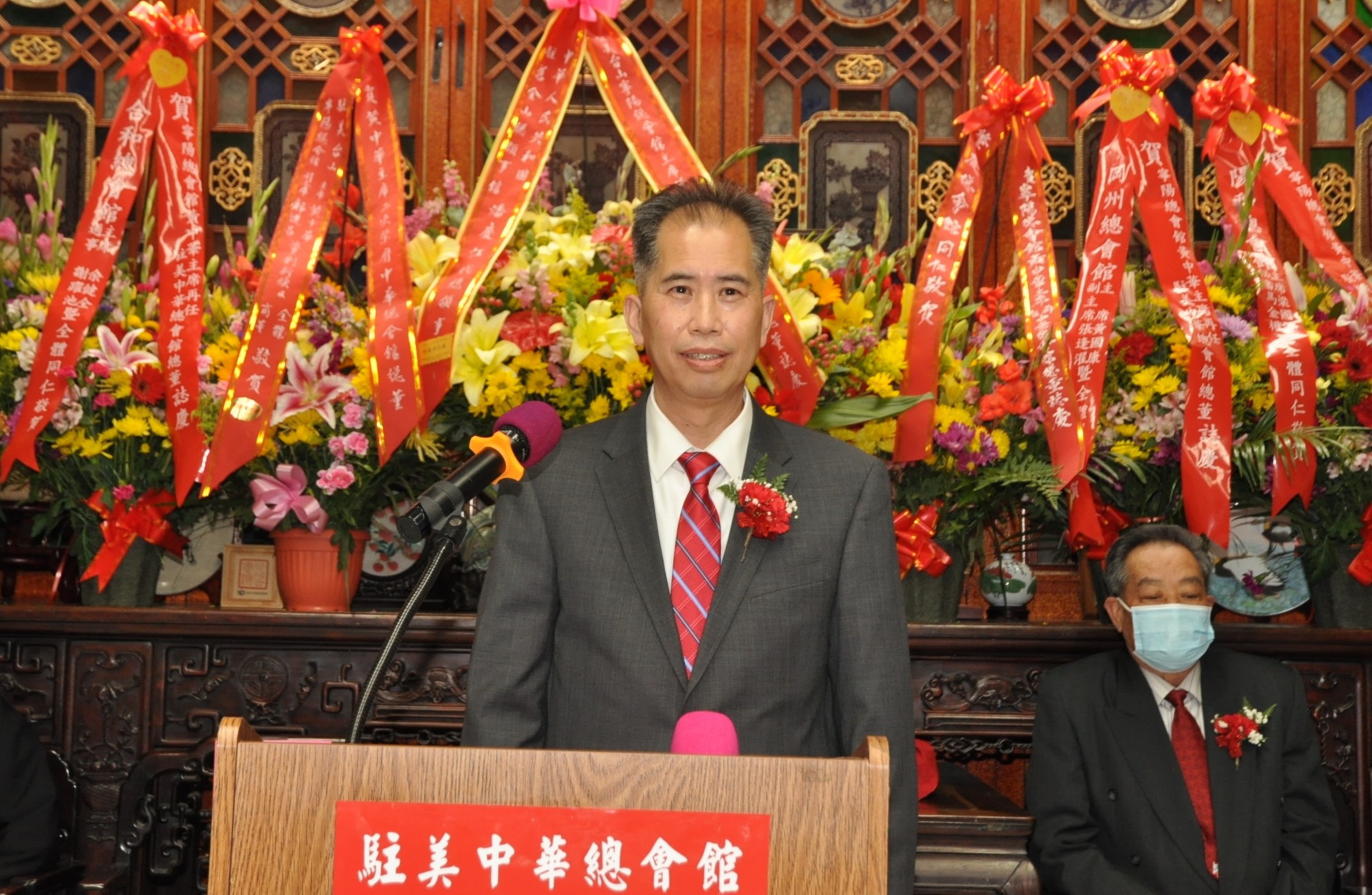 A man in a suit and red tie stands behind a podium with red decorations and floral arrangements behind him, while another man wearing a mask sits nearby.