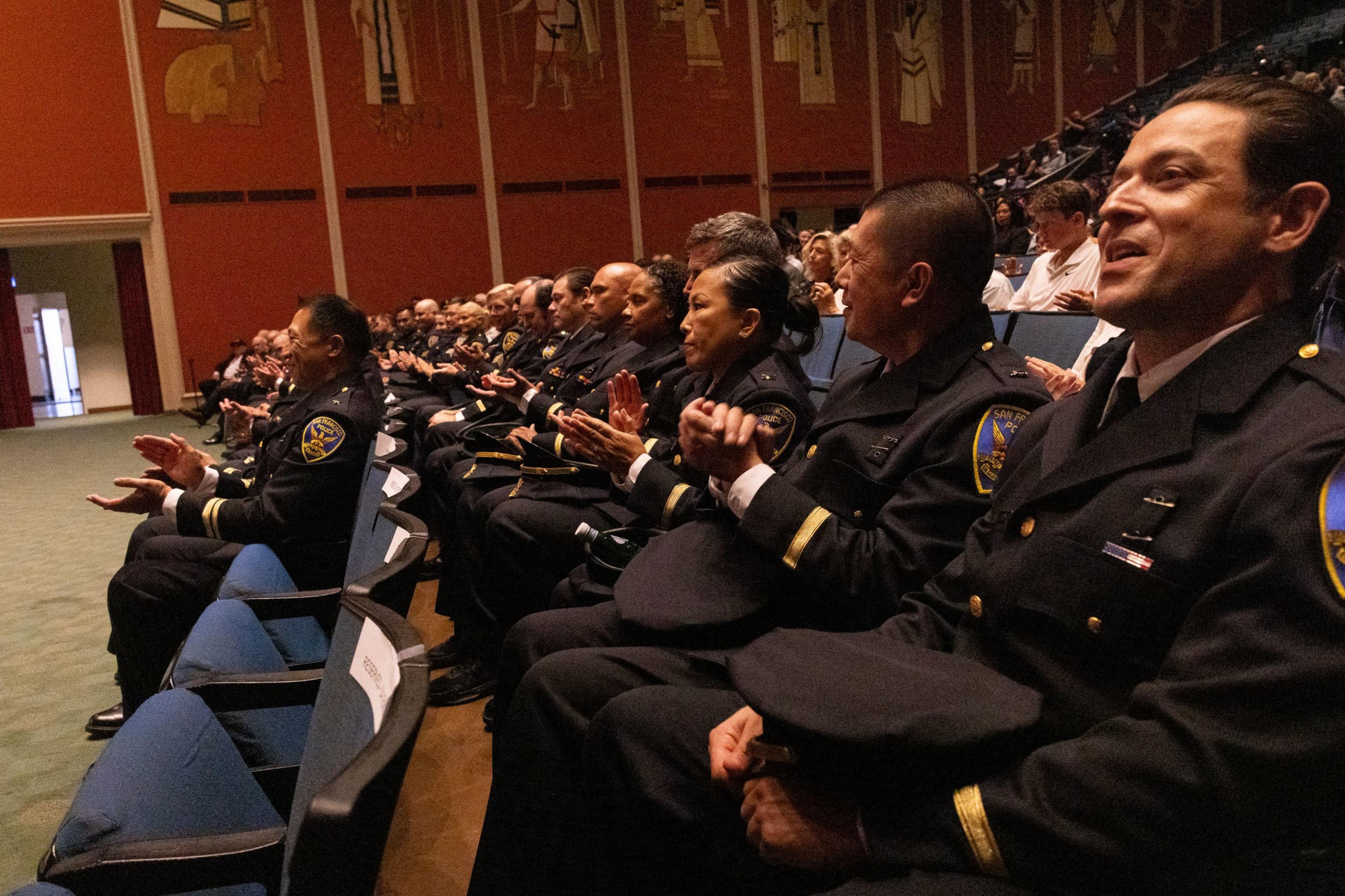 A group of uniformed officers sits in rows of a large auditorium, clapping and engaging attentively, with an ornate, mural-covered wall in the background.