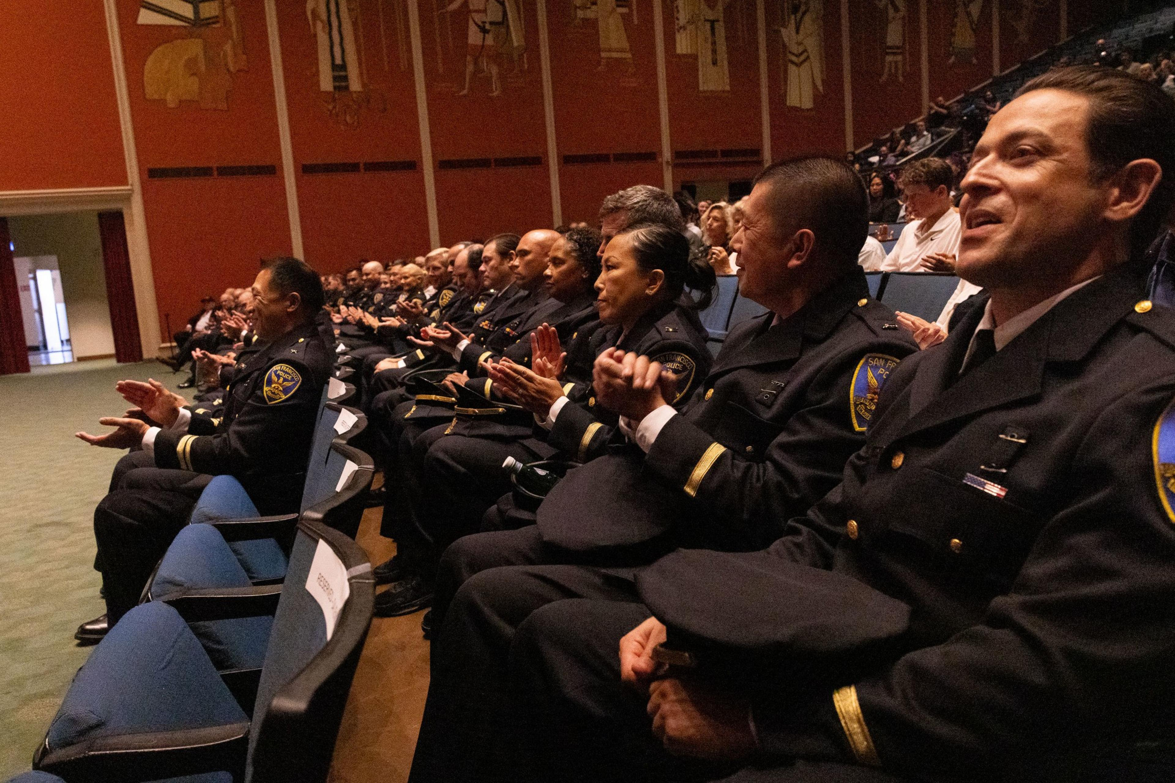 A group of uniformed officers sits in rows of a large auditorium, clapping and engaging attentively, with an ornate, mural-covered wall in the background.