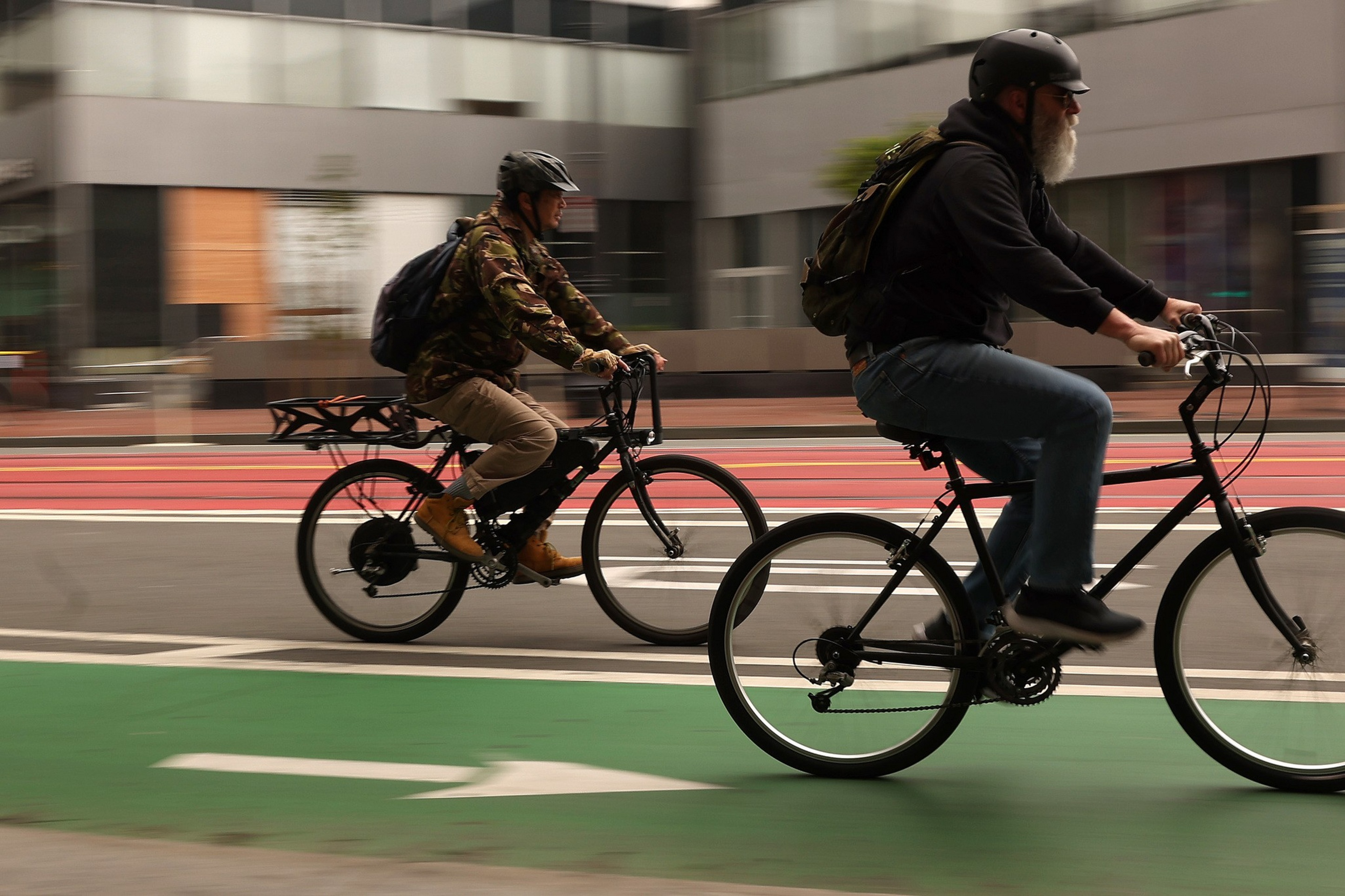 Two men wearing helmets ride bicycles on a green bike lane in an urban area with blurred buildings in the background.