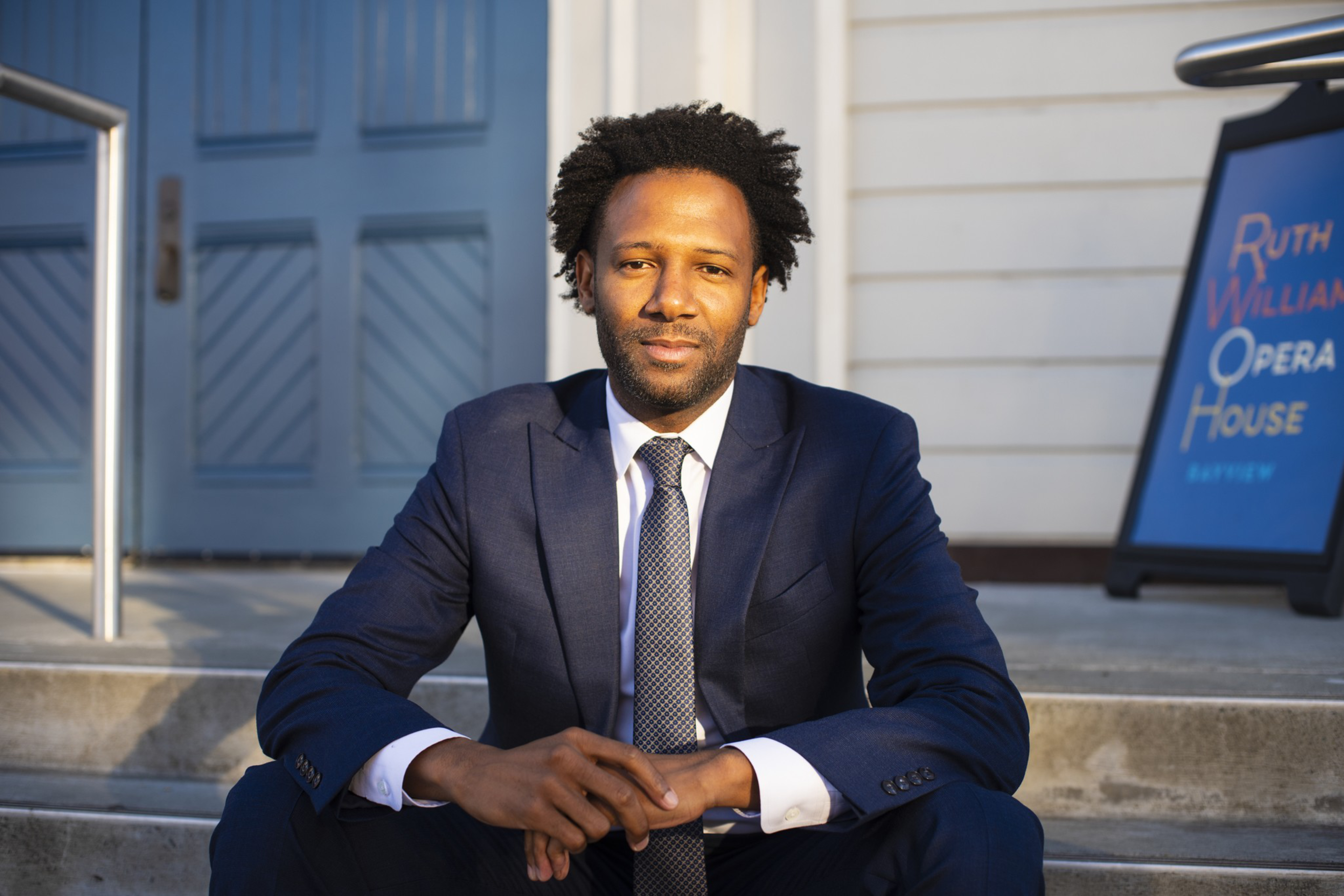A man in a navy blue suit with a white shirt and tie sits on concrete steps outside a building with a blue sign reading “Ruth Williams Opera House.”
