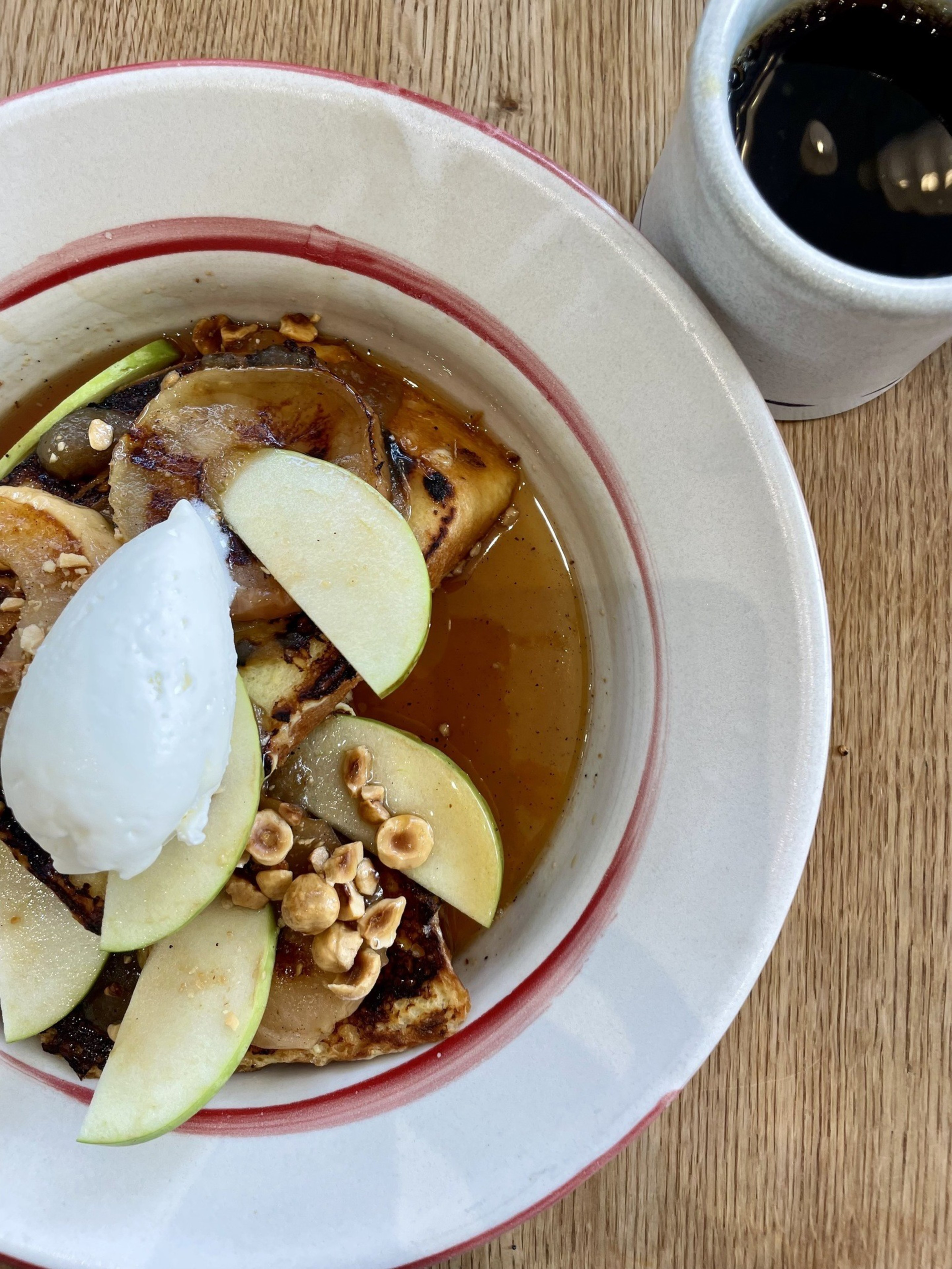 A dish with roasted vegetables, nuts, and green apple slices topped with a scoop of sorbet sits in a white bowl with a red rim. A cup of black coffee is beside it.