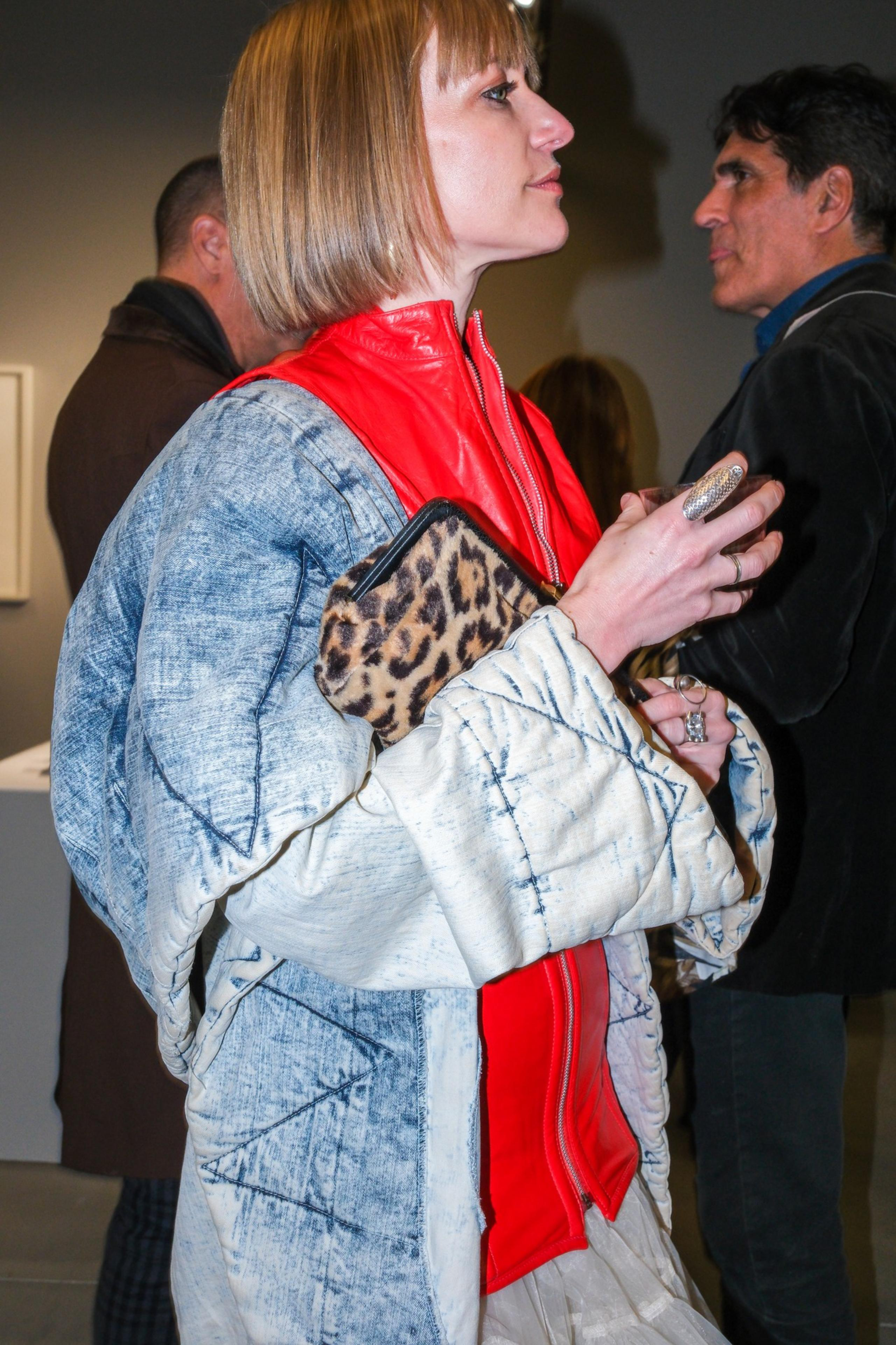 A person with short hair in a red jacket under a blue patterned outer layer holds a leopard print clutch and a drink. Others are in the background.