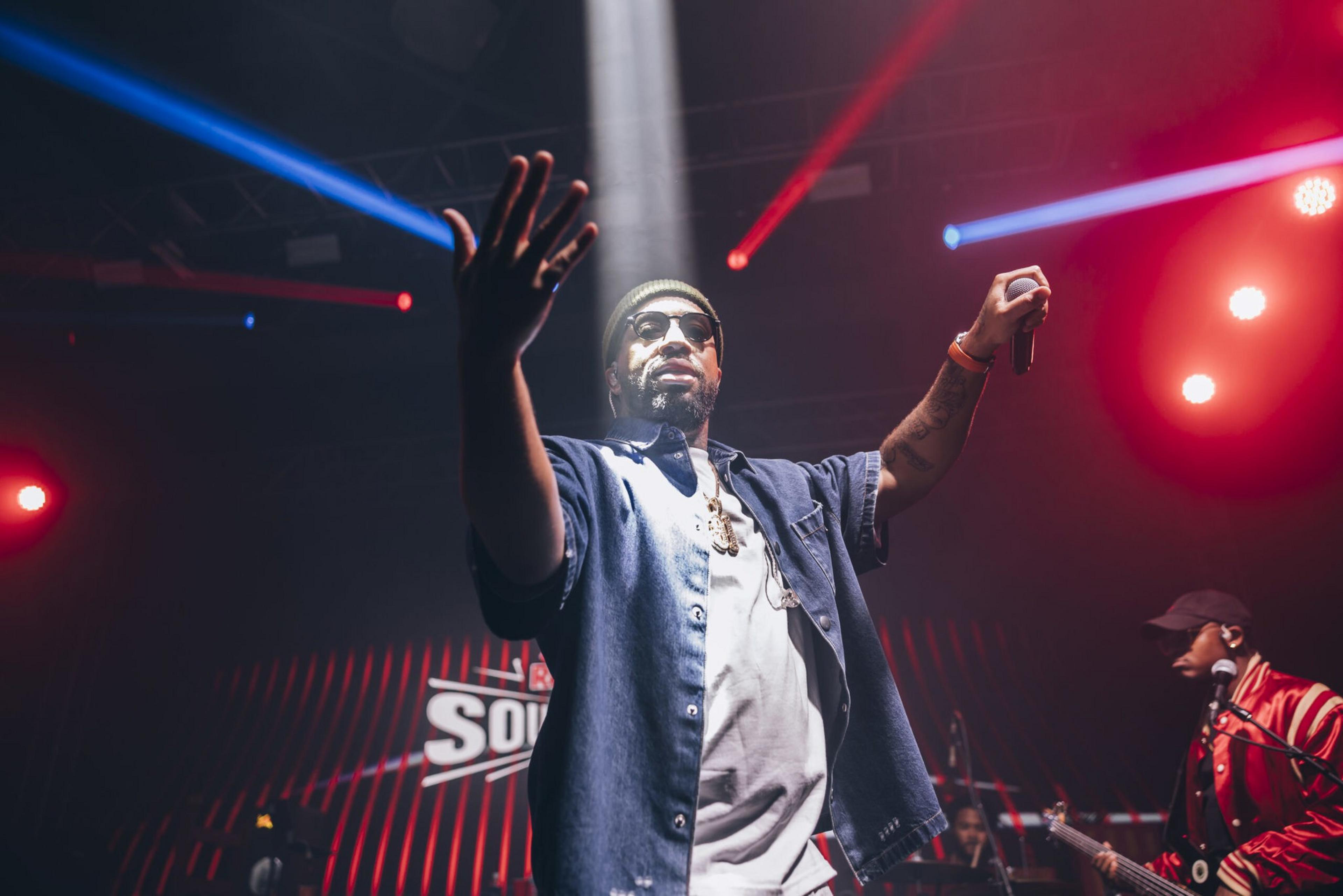 A man wearing sunglasses and a beanie holds a microphone with one hand raised, performing on stage under colorful red and blue lights.