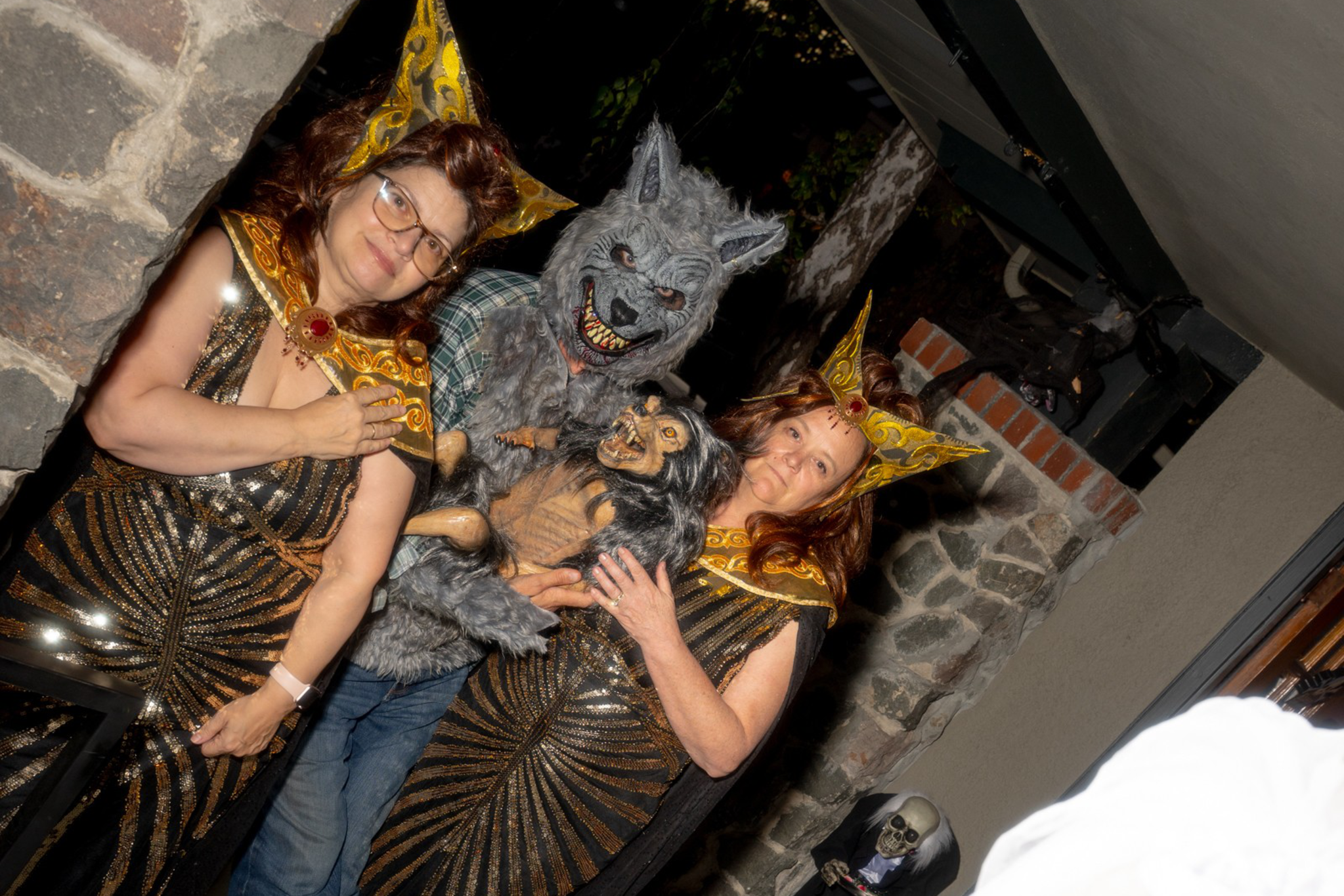 Two women in golden Egyptian-style costumes stand next to a person in a werewolf mask holding a monkey puppet, with a ghostly figure in white blurred in the foreground.
