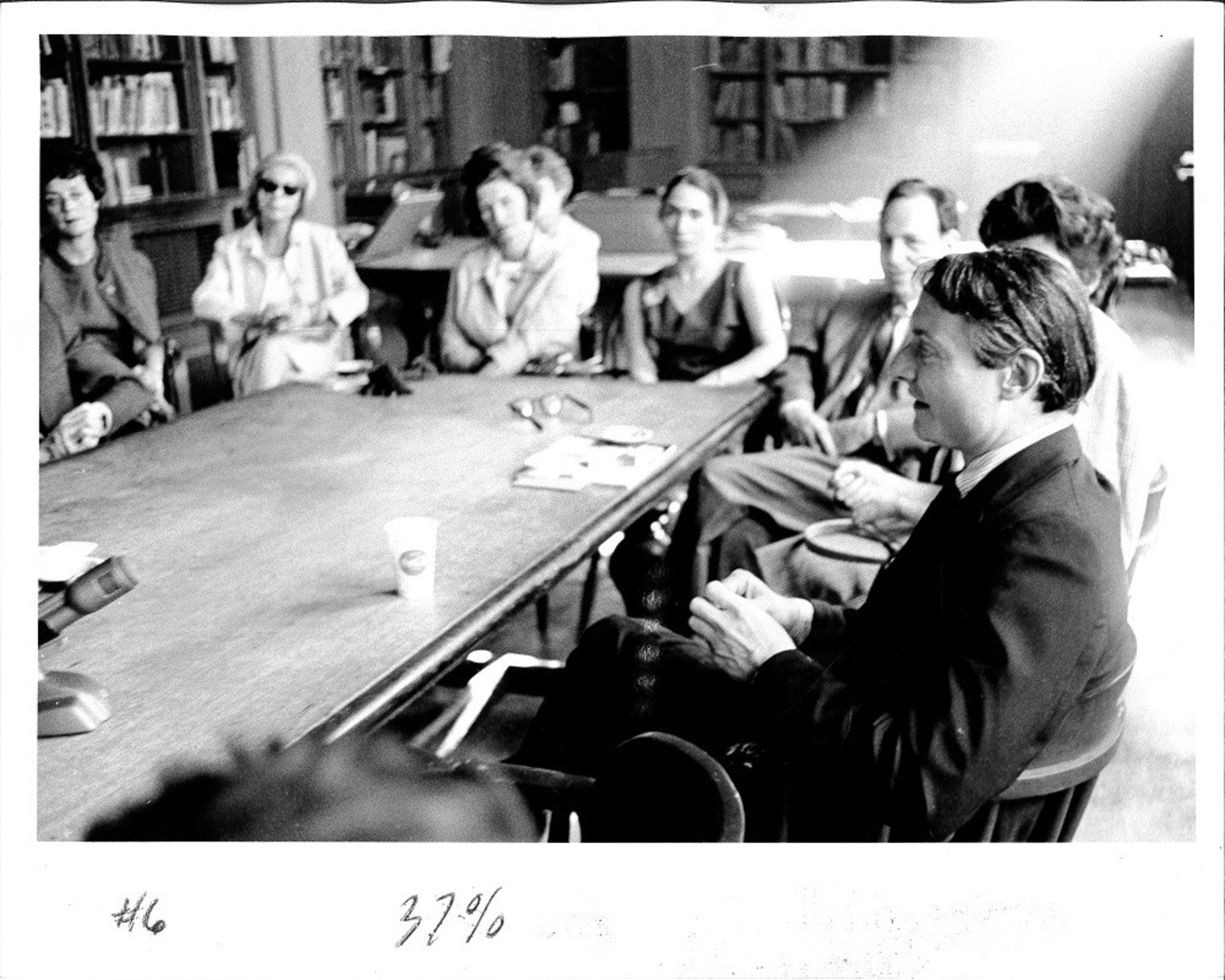 A group of people sits around a long table in a library, engaged in a discussion, with bookshelves visible in the background.