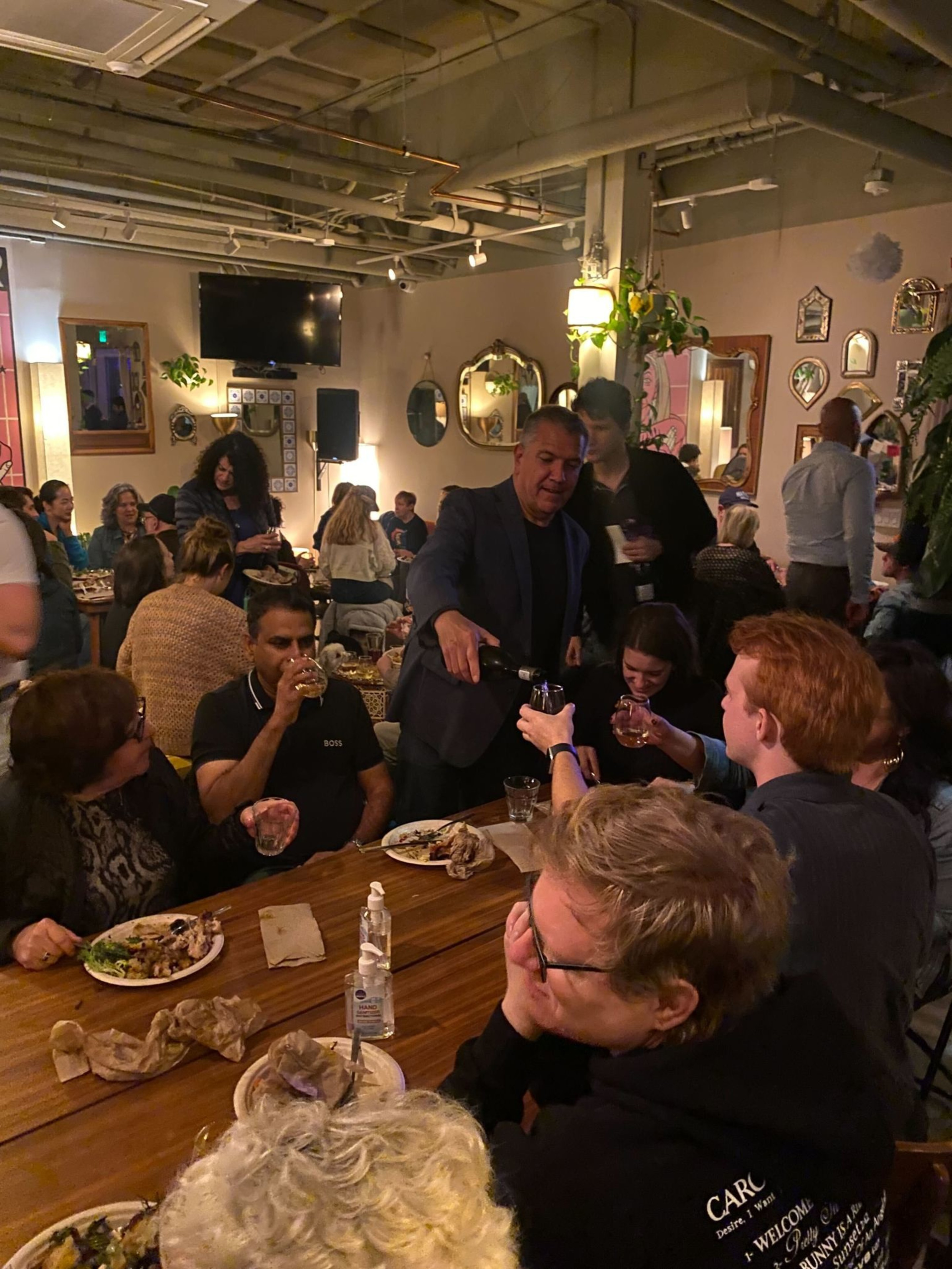 A lively restaurant scene features people seated at a long table, enjoying food and drinks. A man in a suit pours wine, and the room is decorated with mirrors and plants.