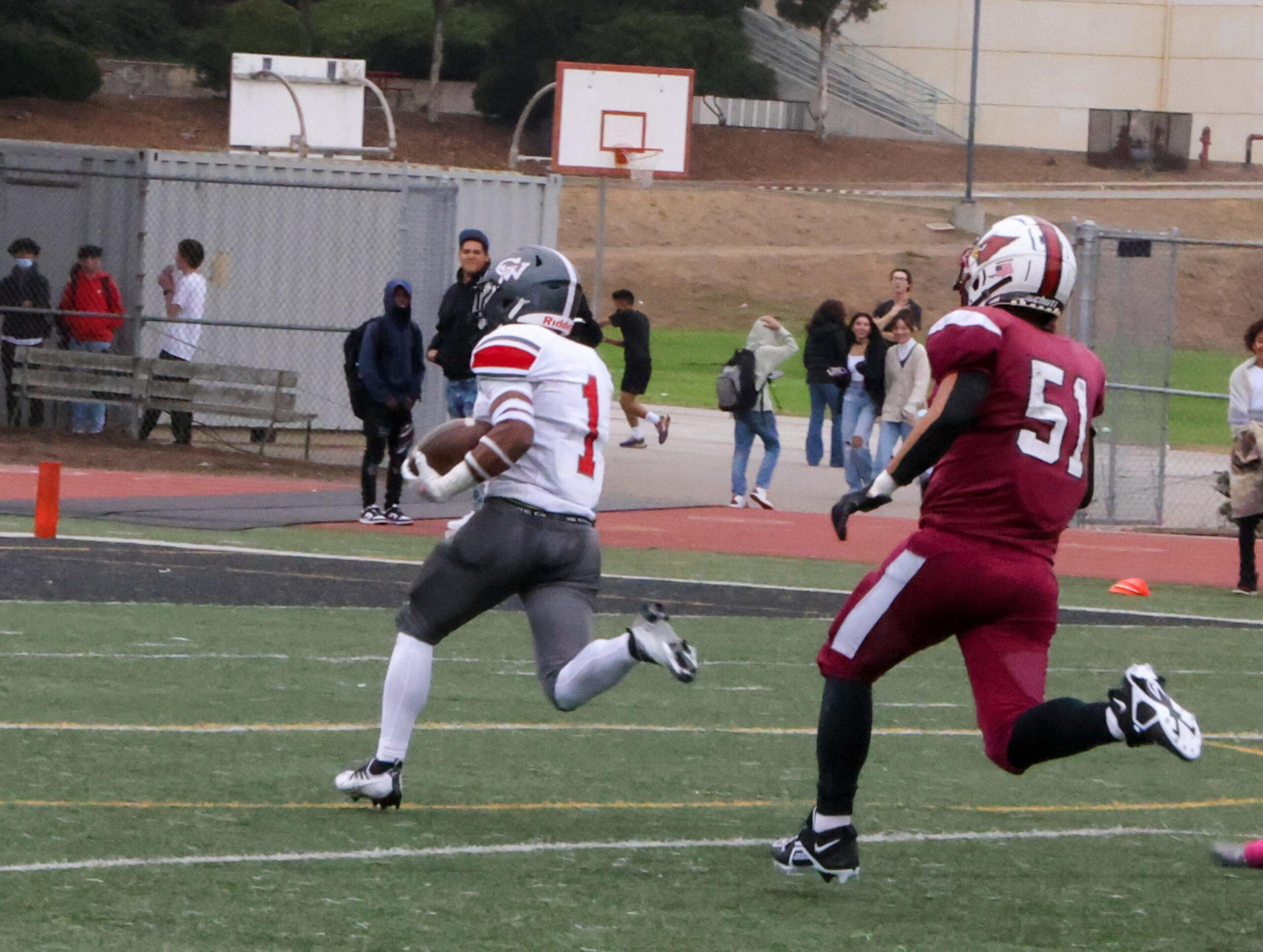 A football player wearing a white and gray uniform with number 1 runs with the ball while a player in a maroon uniform with number 51 chases him.
