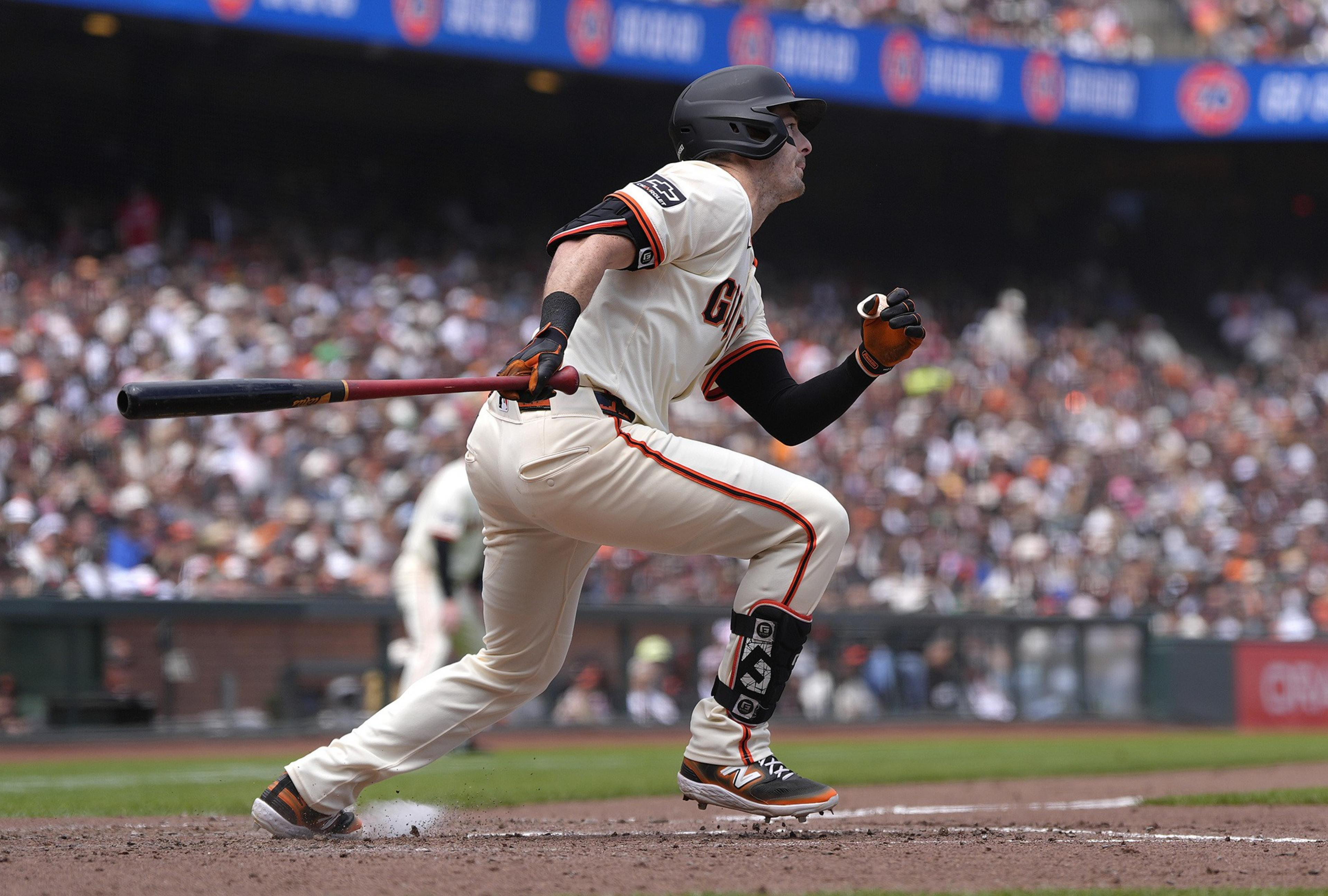 A baseball player in a white and orange uniform is running, gripping a bat. The crowd in the background is blurred, suggesting a lively ballpark scene.