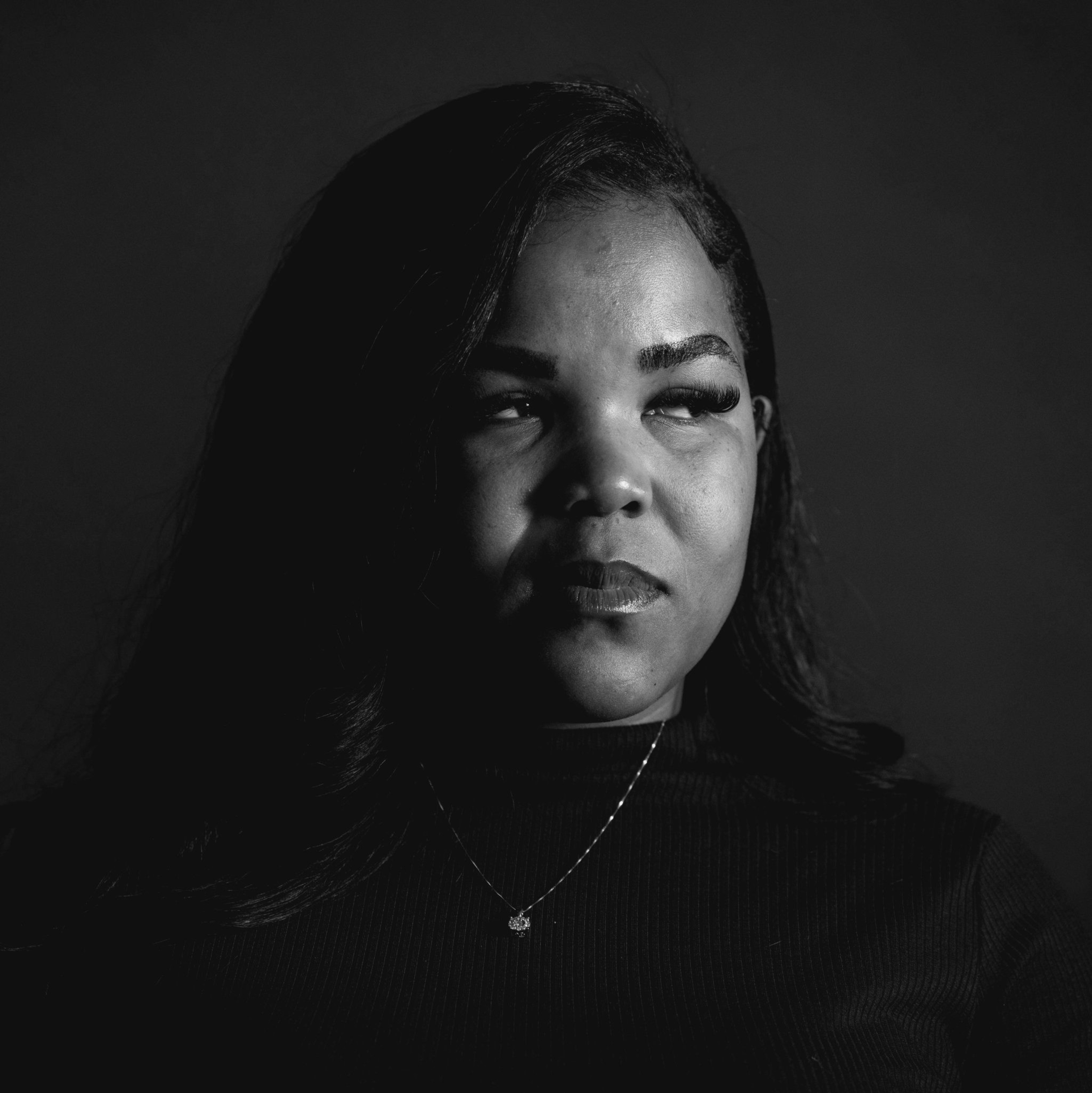 A black and white portrait of a woman gazing upward to the right. A soft light highlights her face and a necklace rests on her dark top.