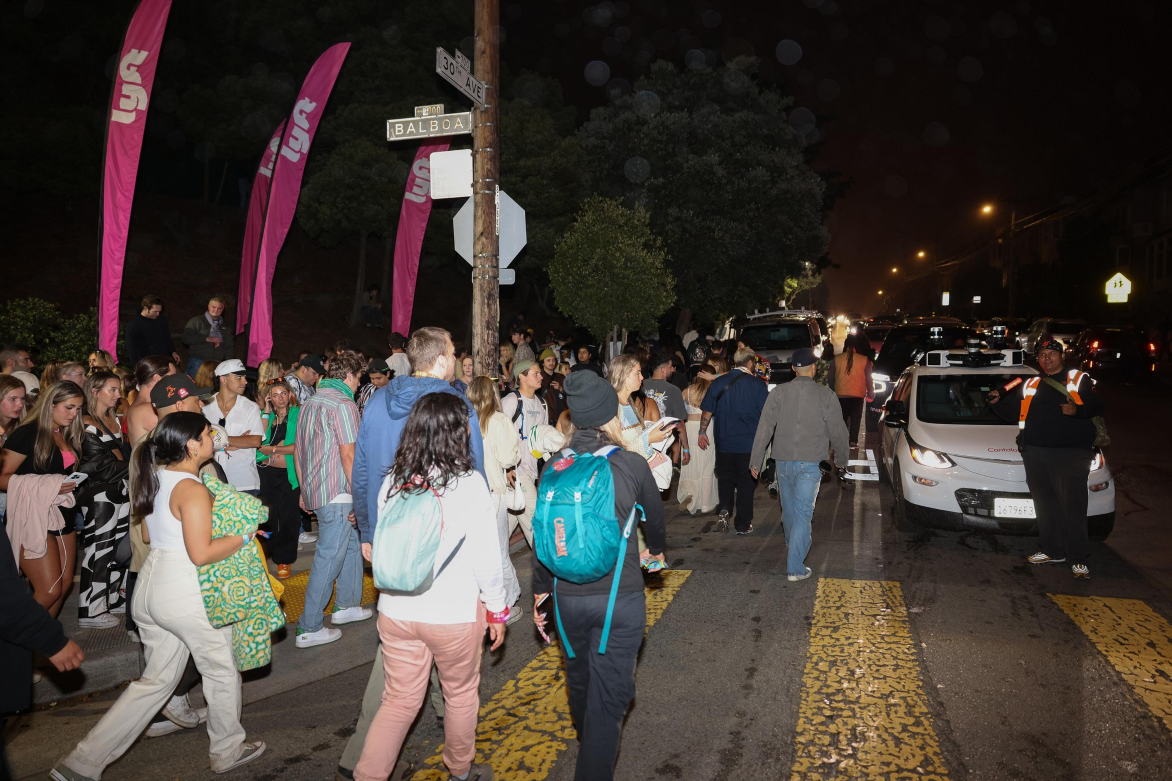 A large group of people waits at a crosswalk on a street corner at night, near pink Lyft flags and a white Lyft car with an attendant.