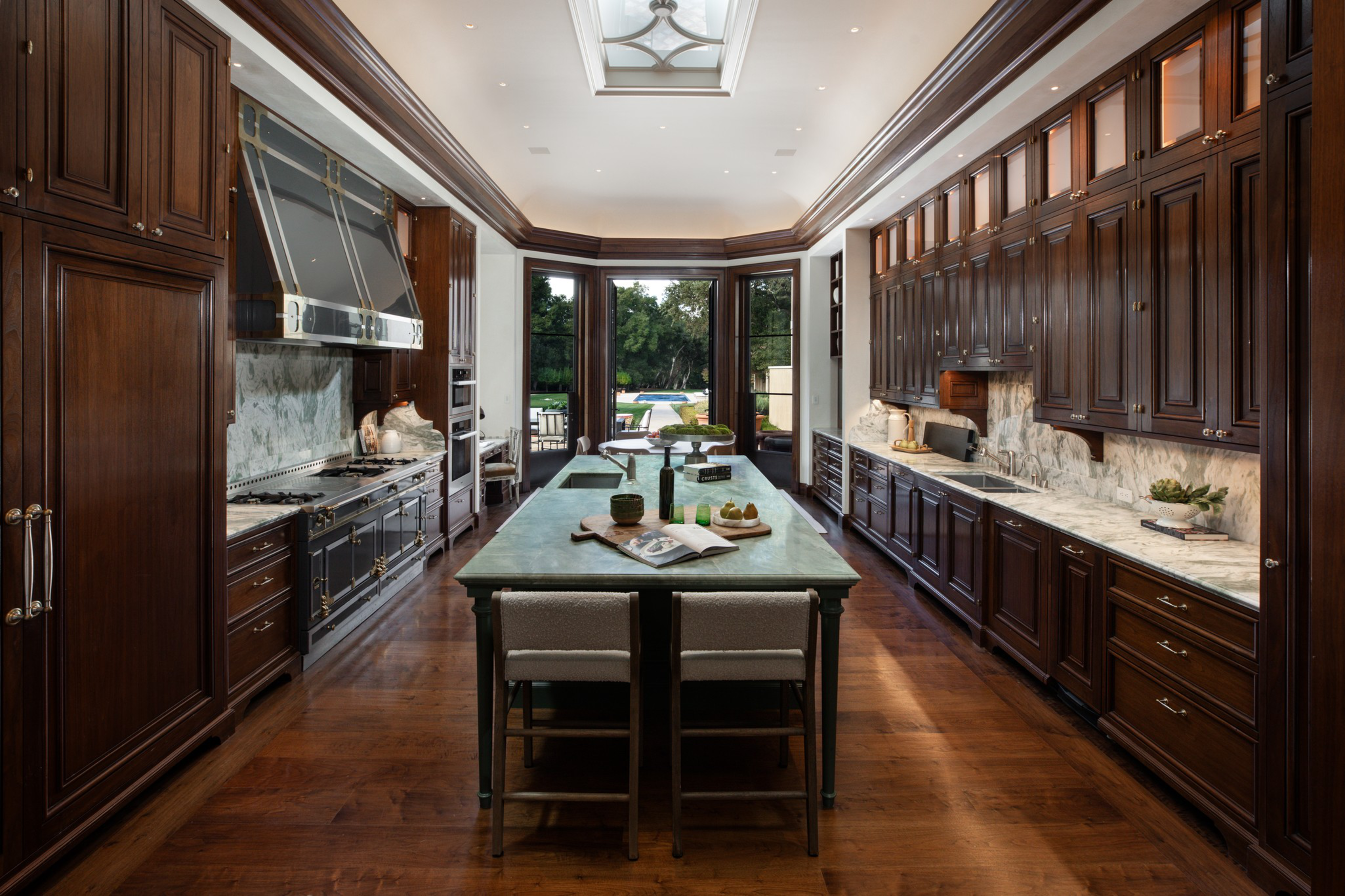 A spacious kitchen features dark wood cabinets, a long central island with seating, marble countertops, stainless appliances, and large windows overlooking a garden.