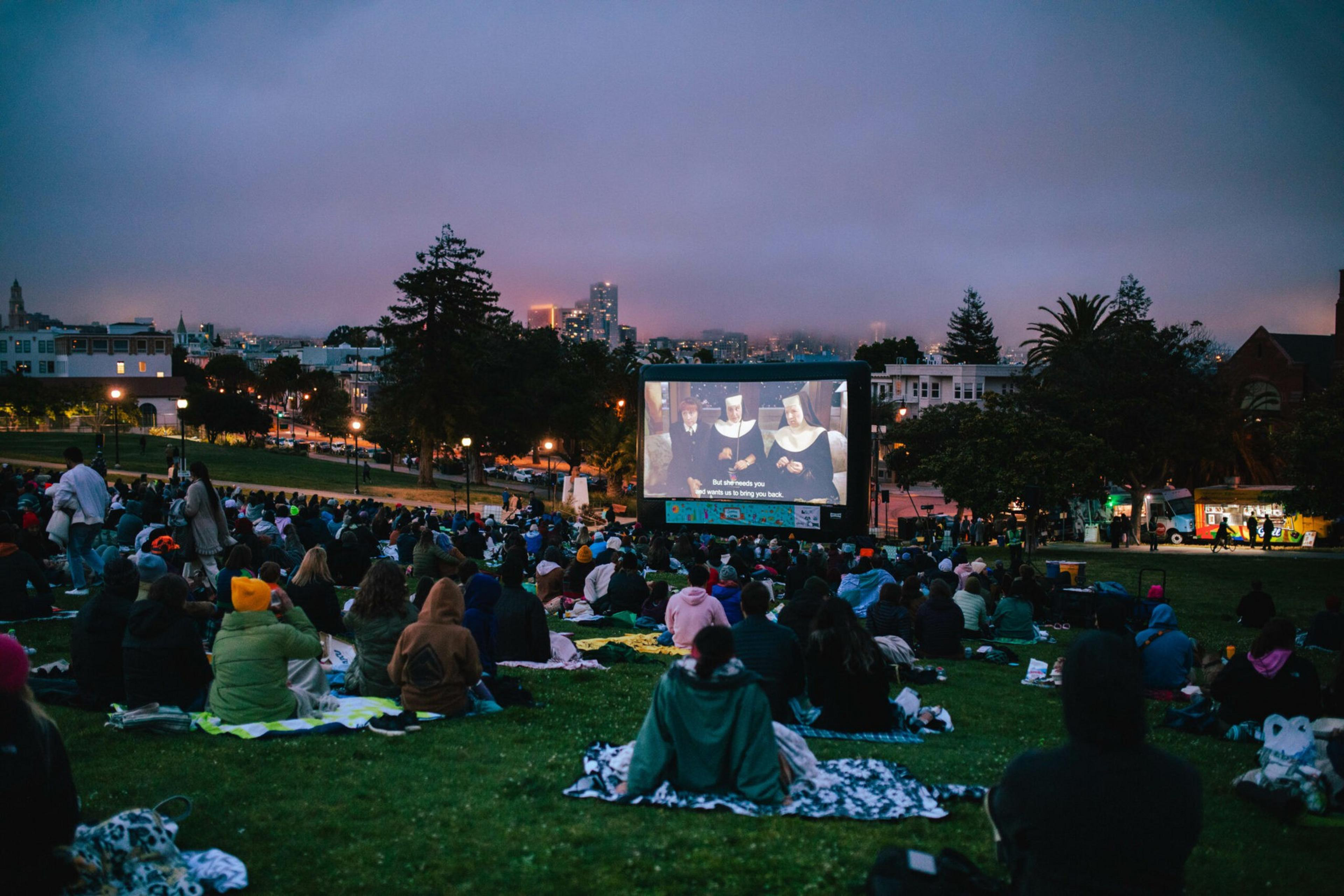 A crowd sits on blankets and benches in a park at dusk, watching a movie on a large outdoor screen, with city buildings and trees in the background.