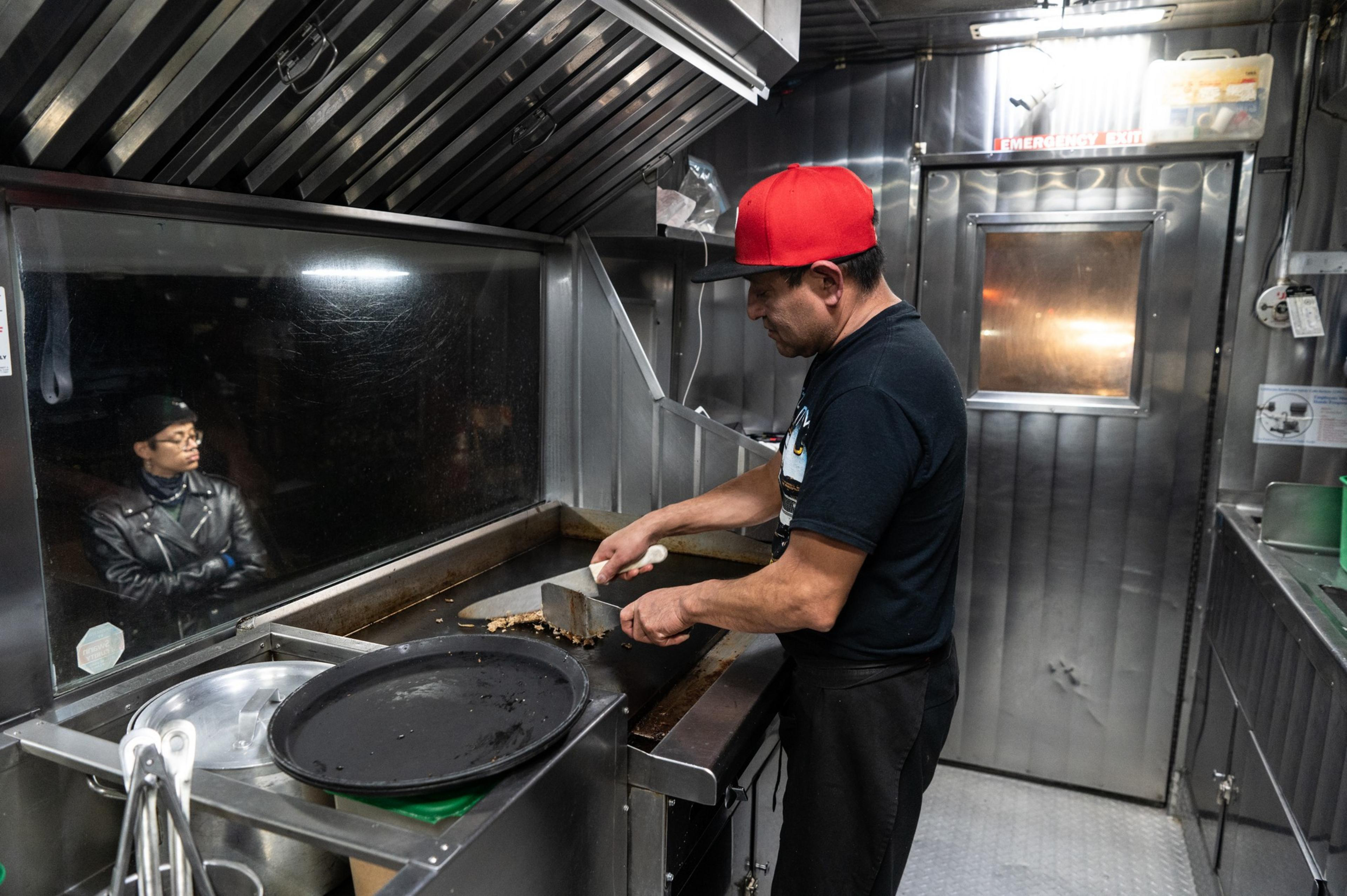 A man in a red cap cooks on a griddle inside a food truck. A reflection of a person wearing glasses and a leather jacket is visible in the window.