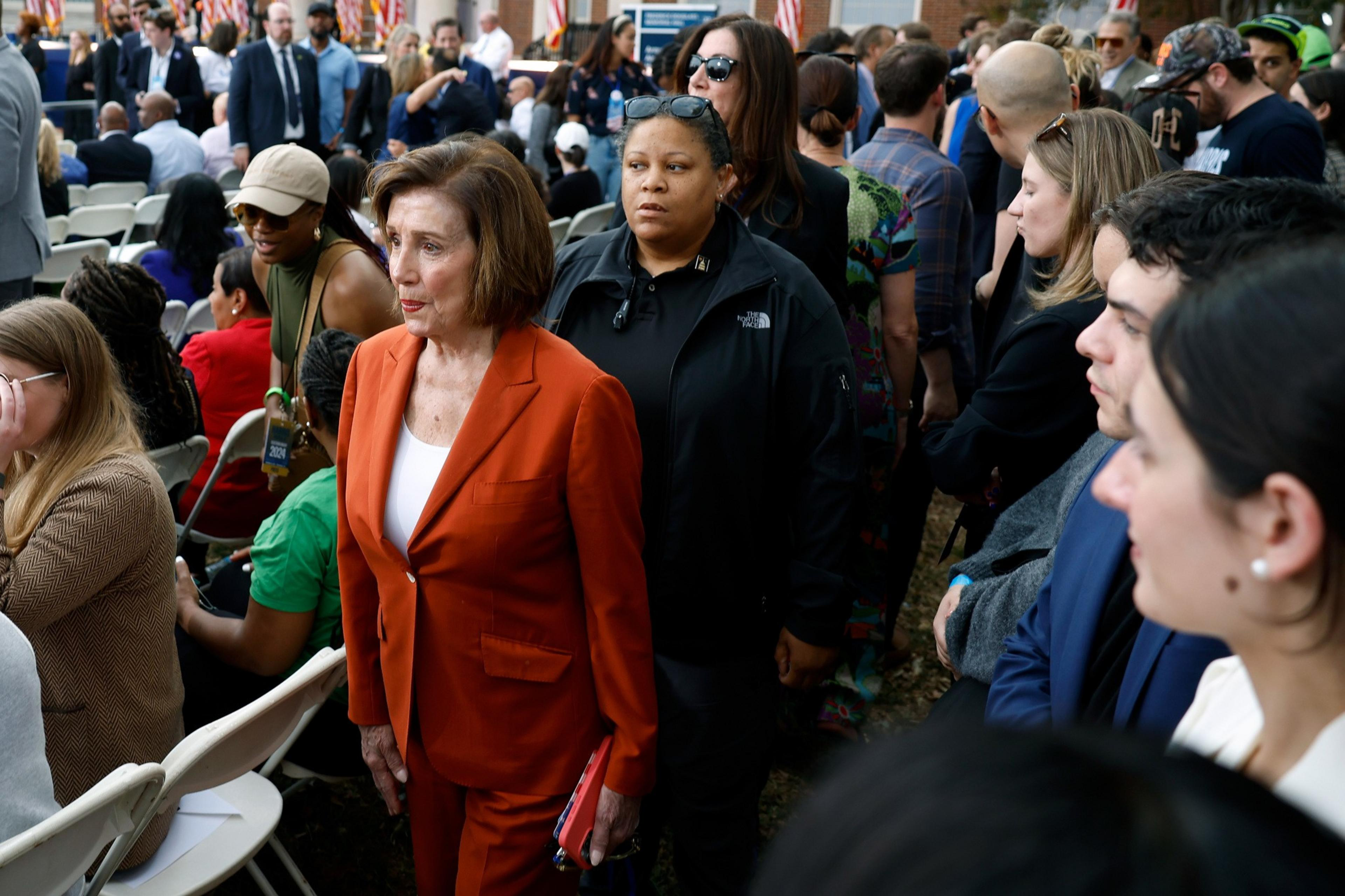A woman in an orange suit stands amidst a crowd seated outdoors, with people talking and walking around. In the background, there are American flags.