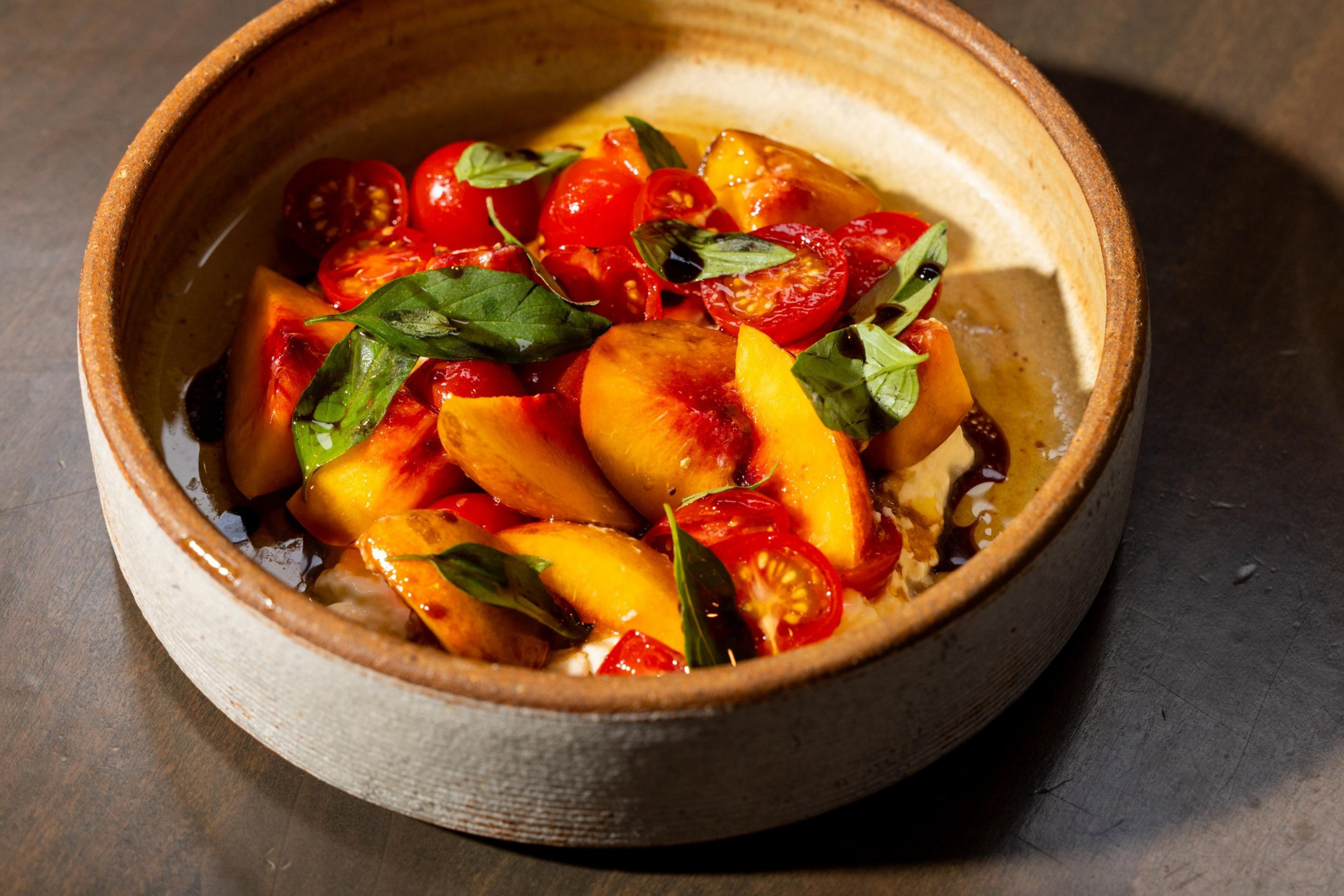 Sliced peaches and cherry tomatoes topped with fresh basil leaves and drizzled with balsamic glaze in a rustic ceramic bowl.