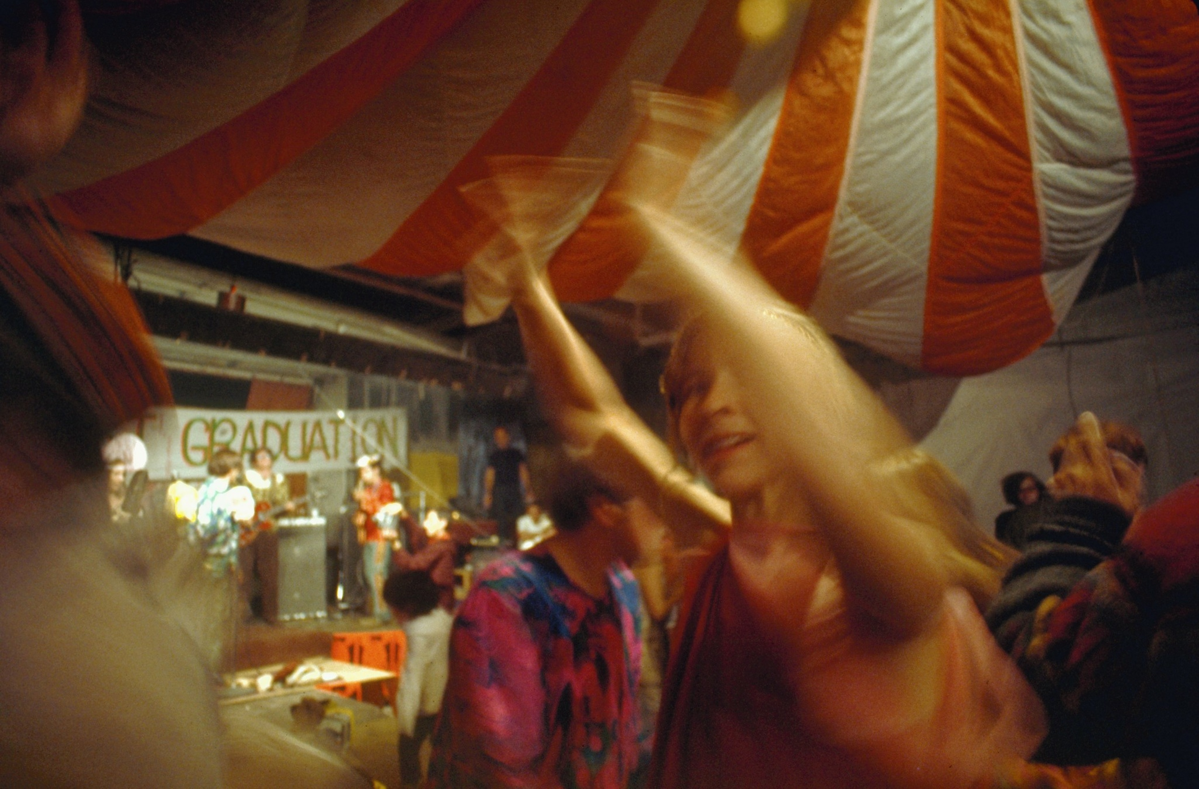 People are dancing energetically under a large red and white striped tent, while a band plays on a stage labeled “Graduation.”