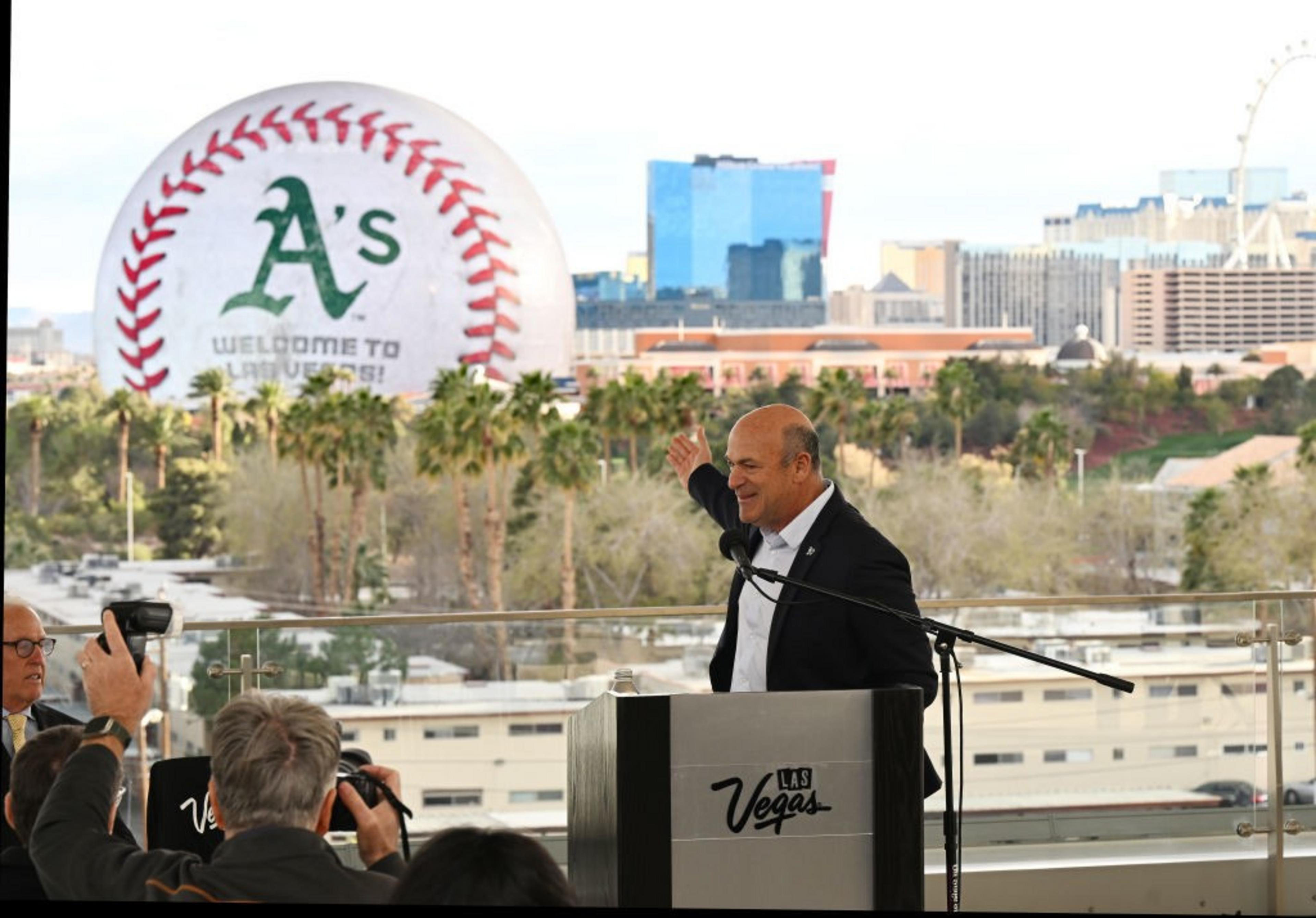A man stands at a podium labeled 'Las Vegas,' addressing an audience. Behind him, a giant baseball with &quot;A's Welcome to Las Vegas!&quot; is visible, set against a cityscape.