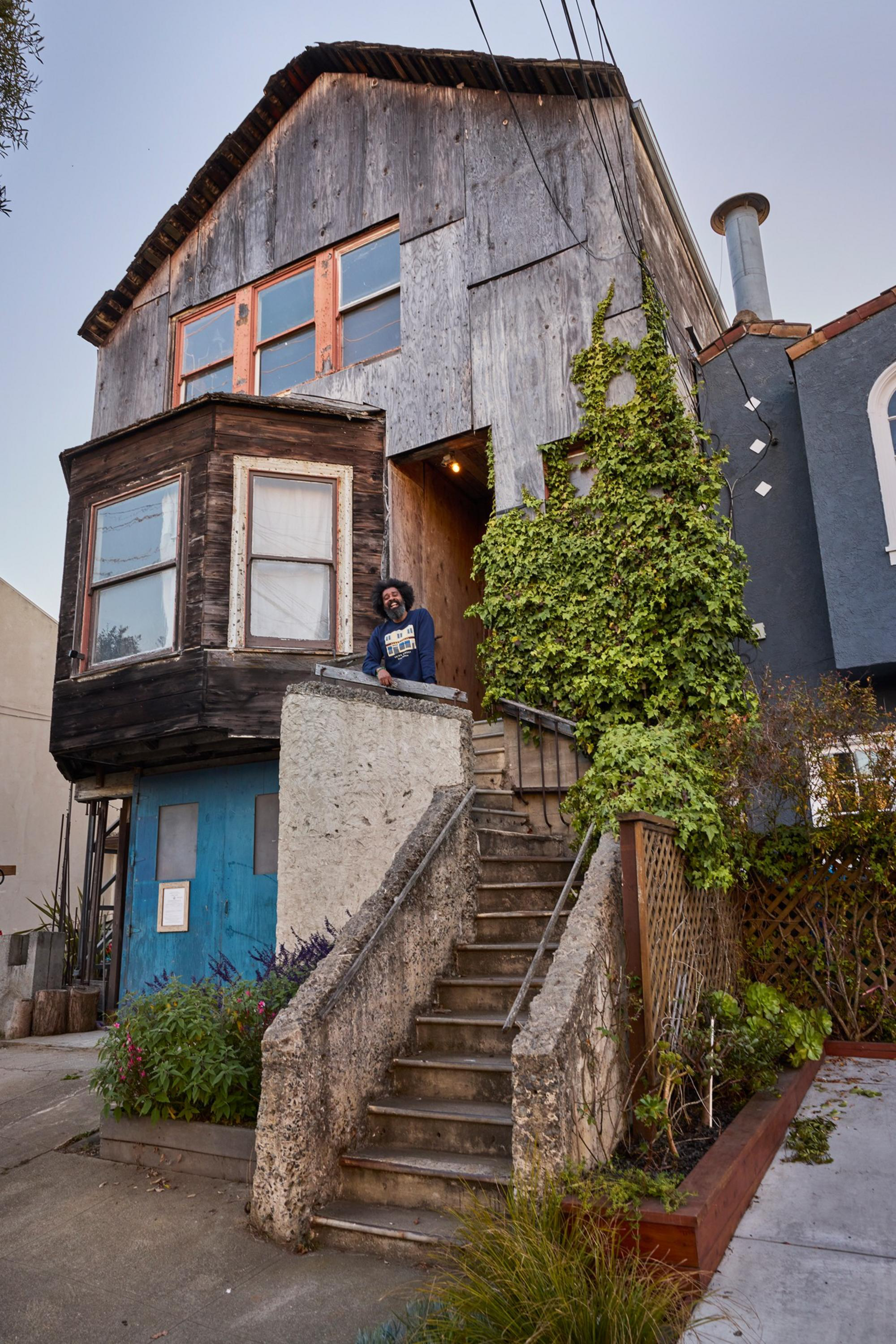 A rustic two-story house features weathered wood beams, a bay window, and a blue door; a person stands on the porch next to tall concrete stairs with greenery.