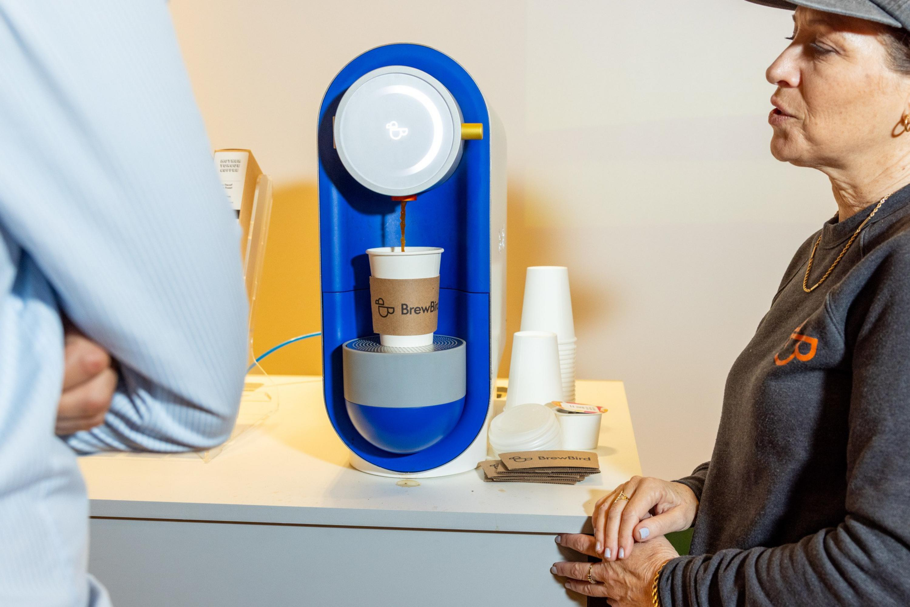 A blue coffee brewing machine is dispensing coffee into a cup labeled "BrewBrew," with two people standing nearby watching.