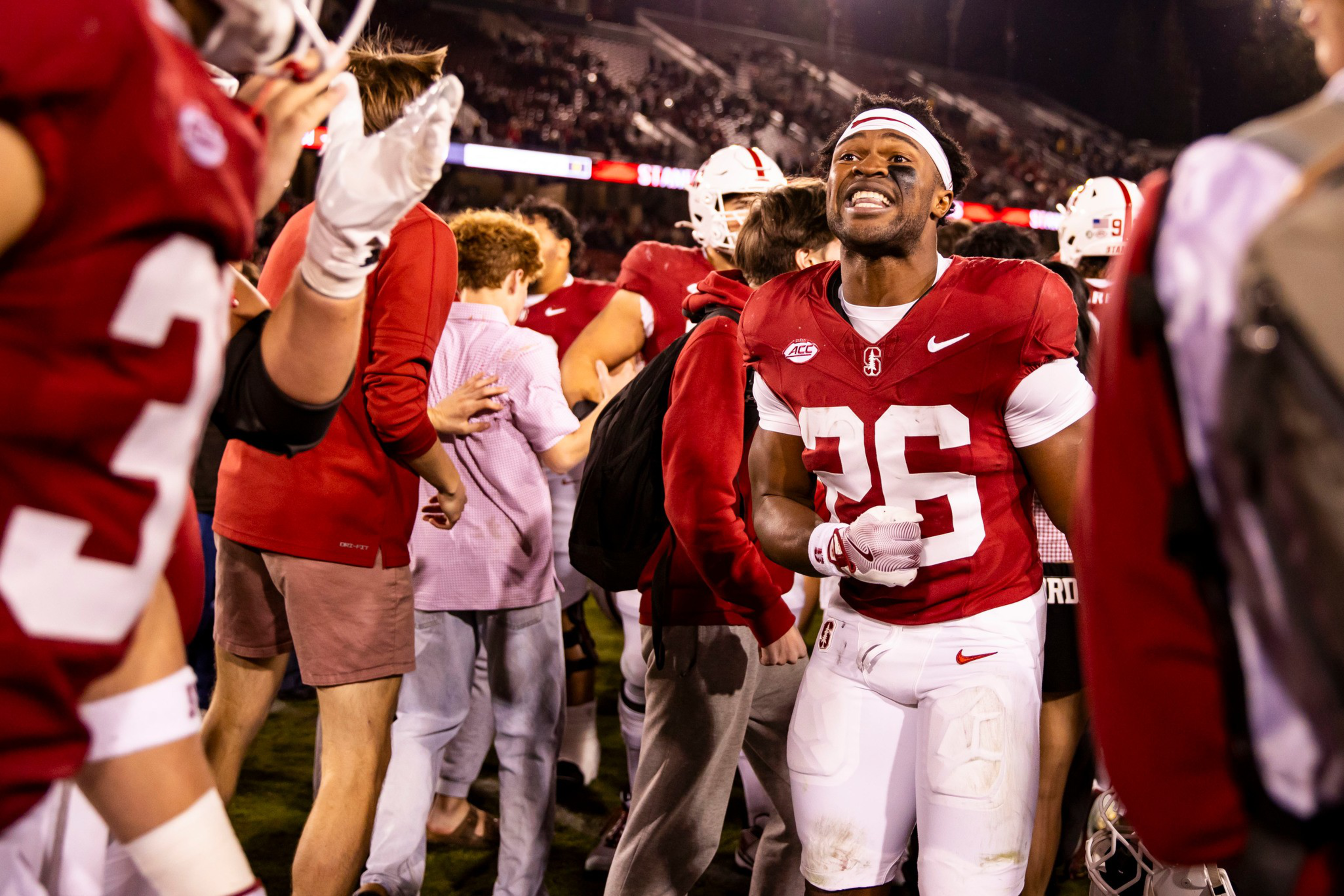 A football player wearing a red jersey with number 26 celebrates on the field among teammates and fans during a night game.