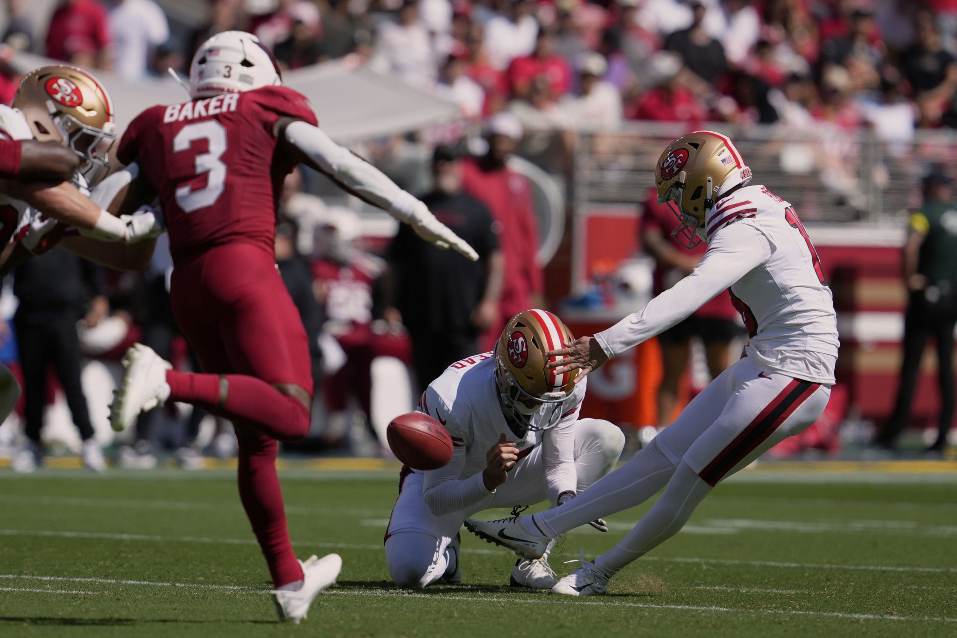 A football player in white kicks the ball held by a teammate while a player in red attempts to block during a game.