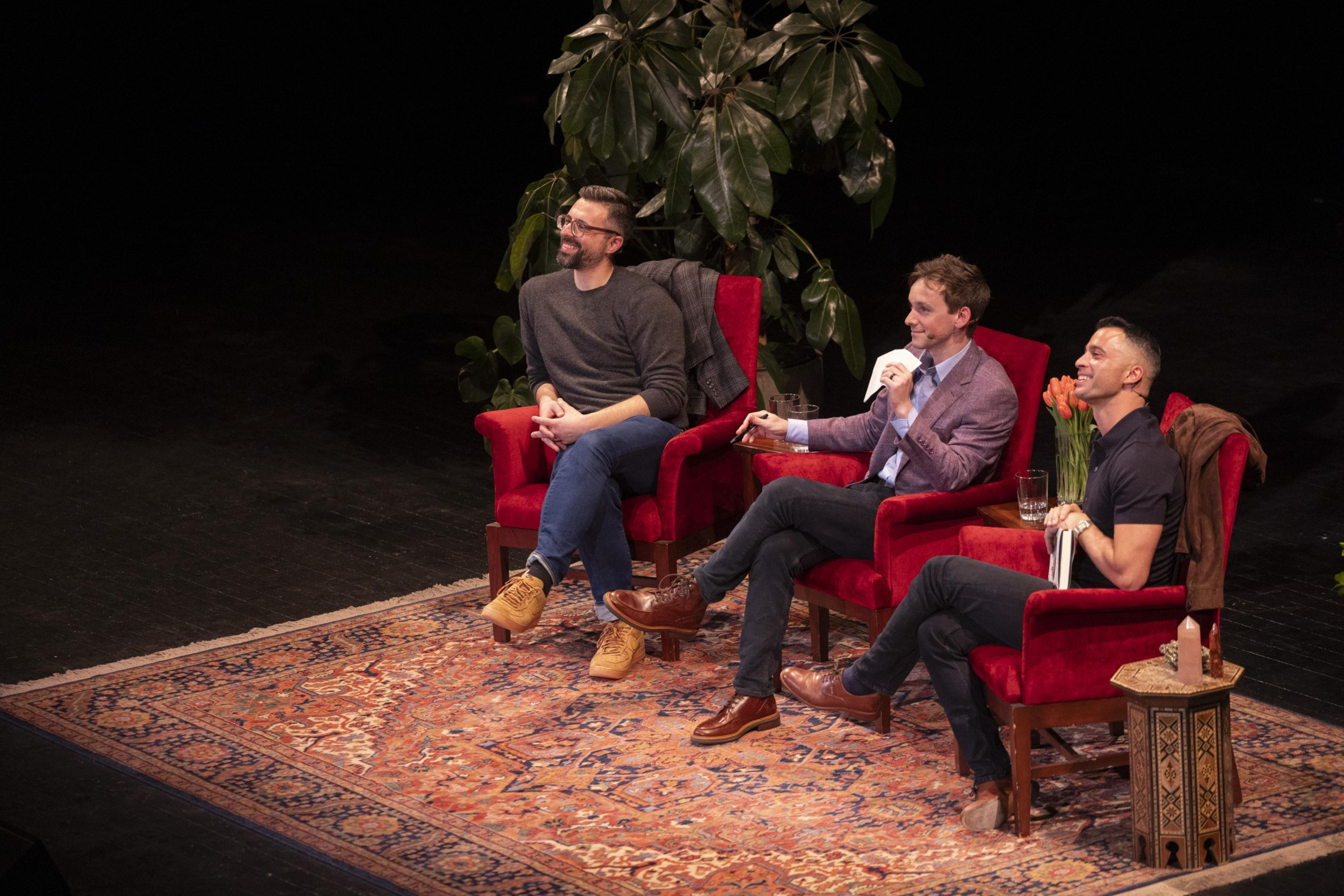 Three people sit on a stage in red armchairs, surrounded by plants and dÃ©cor. They are smiling, engaged in conversation, with a cozy, intimate setting.