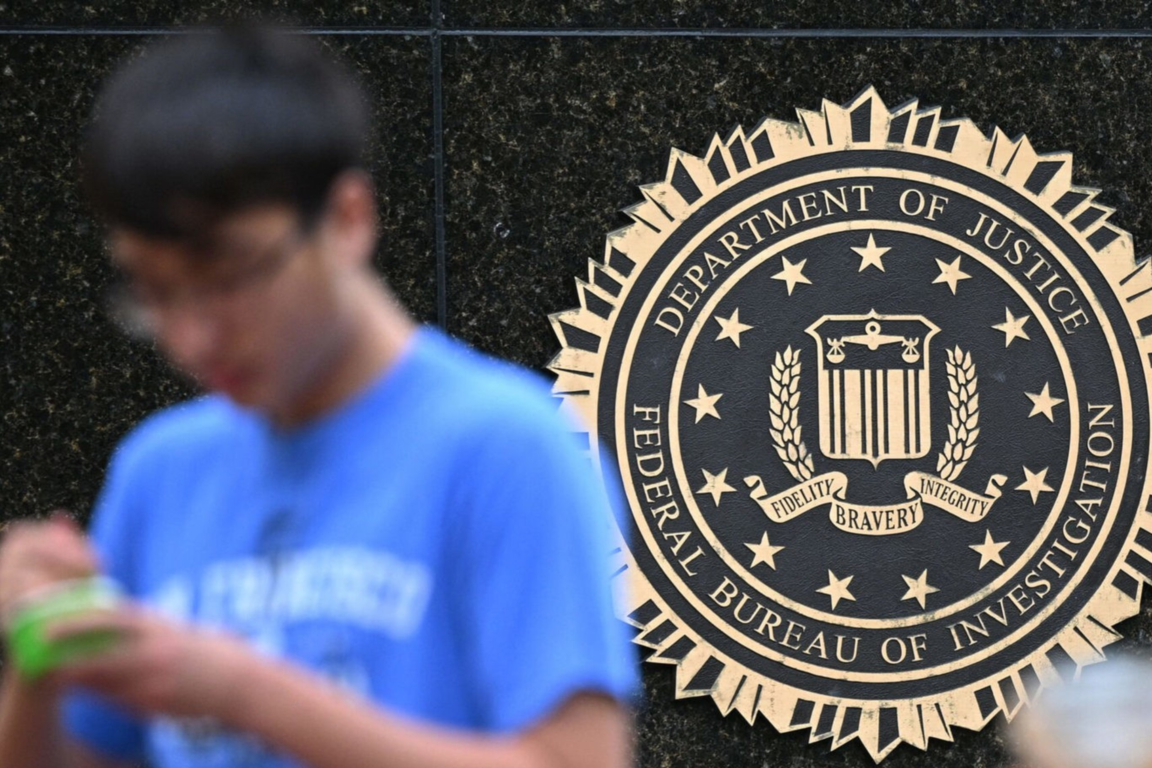 The image shows the FBI seal on a building facade, featuring the Department of Justice emblem. Two blurred figures are in the foreground, one holding a drink.