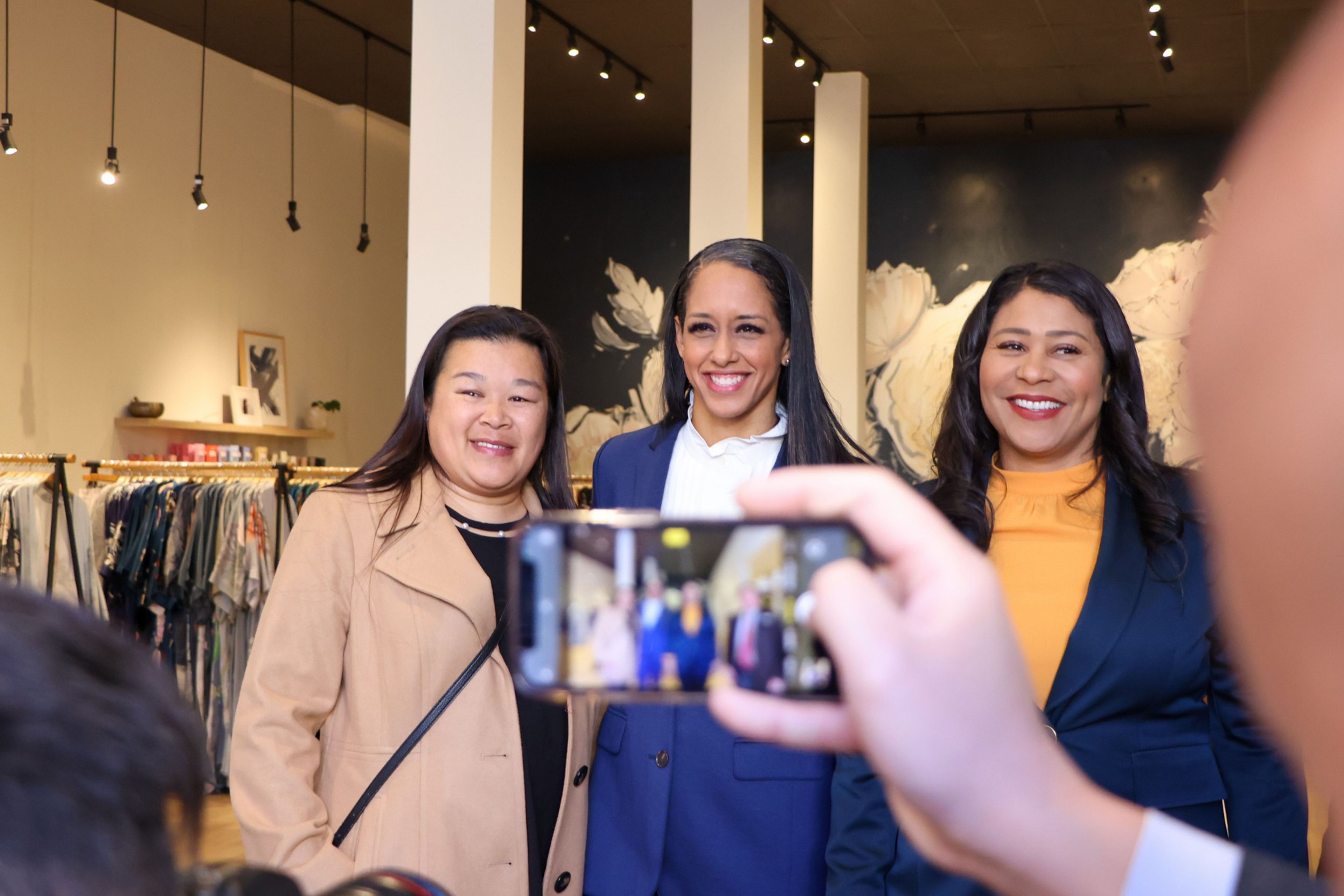 Three smiling women stand closely together inside a clothing store while someone holds a smartphone to take their photo.