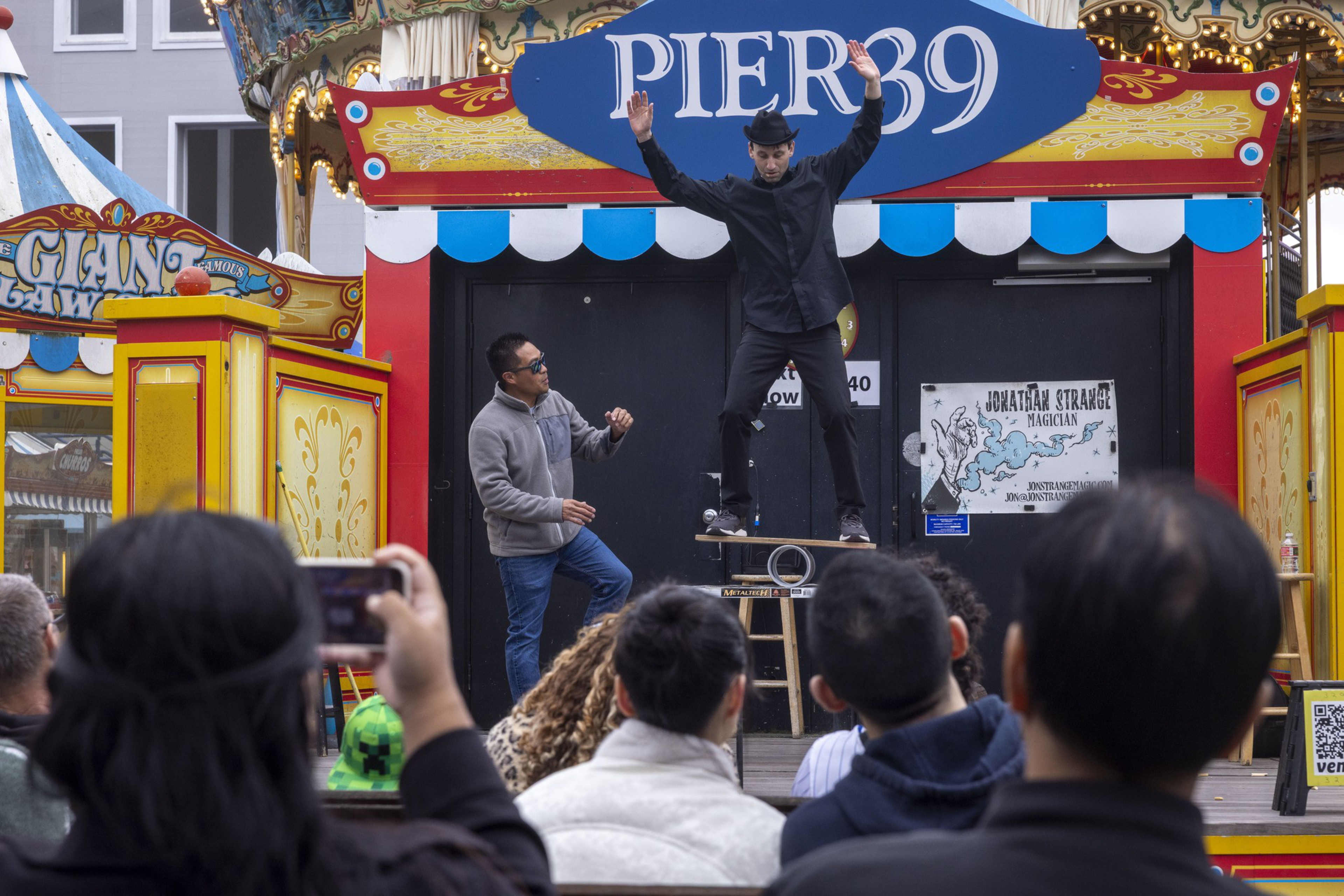 A performer balances on a board atop a cylinder at Pier 39, with a crowd watching. The setting is colorful, with carnival elements in the background.