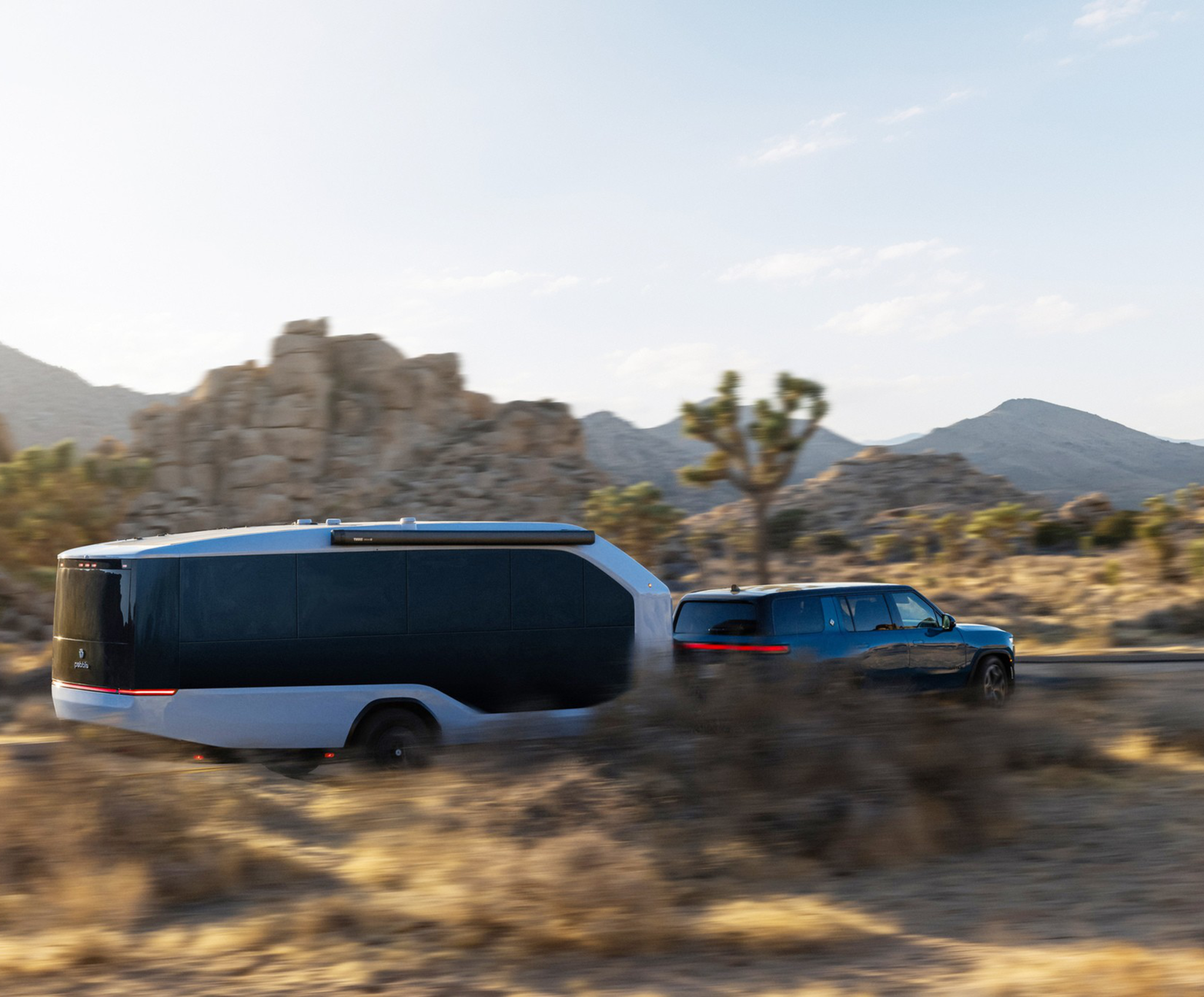A blue SUV is towing a sleek, modern trailer through a desert landscape with rocky hills and sparse vegetation.