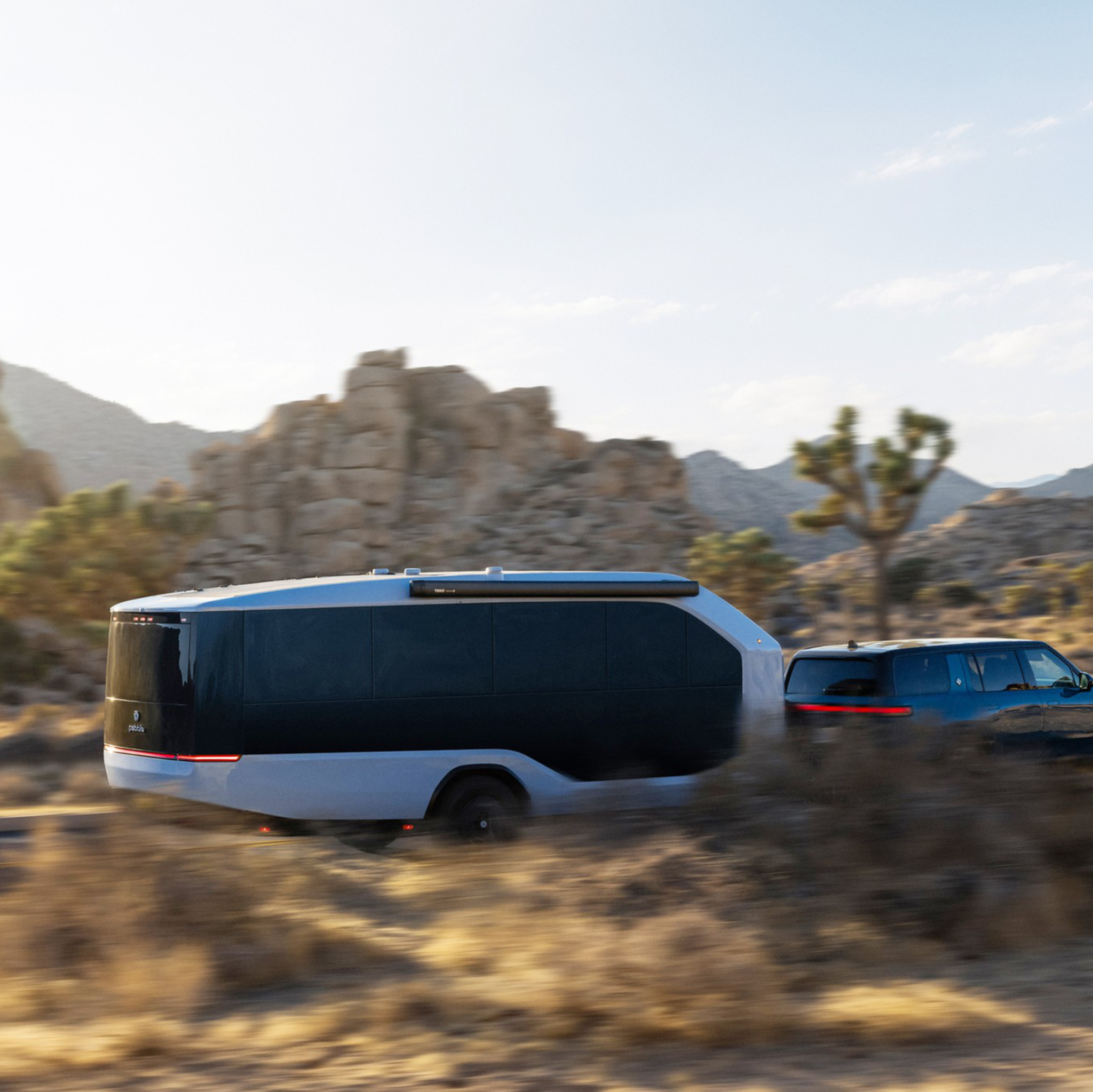 A blue SUV is towing a sleek, modern trailer through a desert landscape with rocky hills and sparse vegetation.