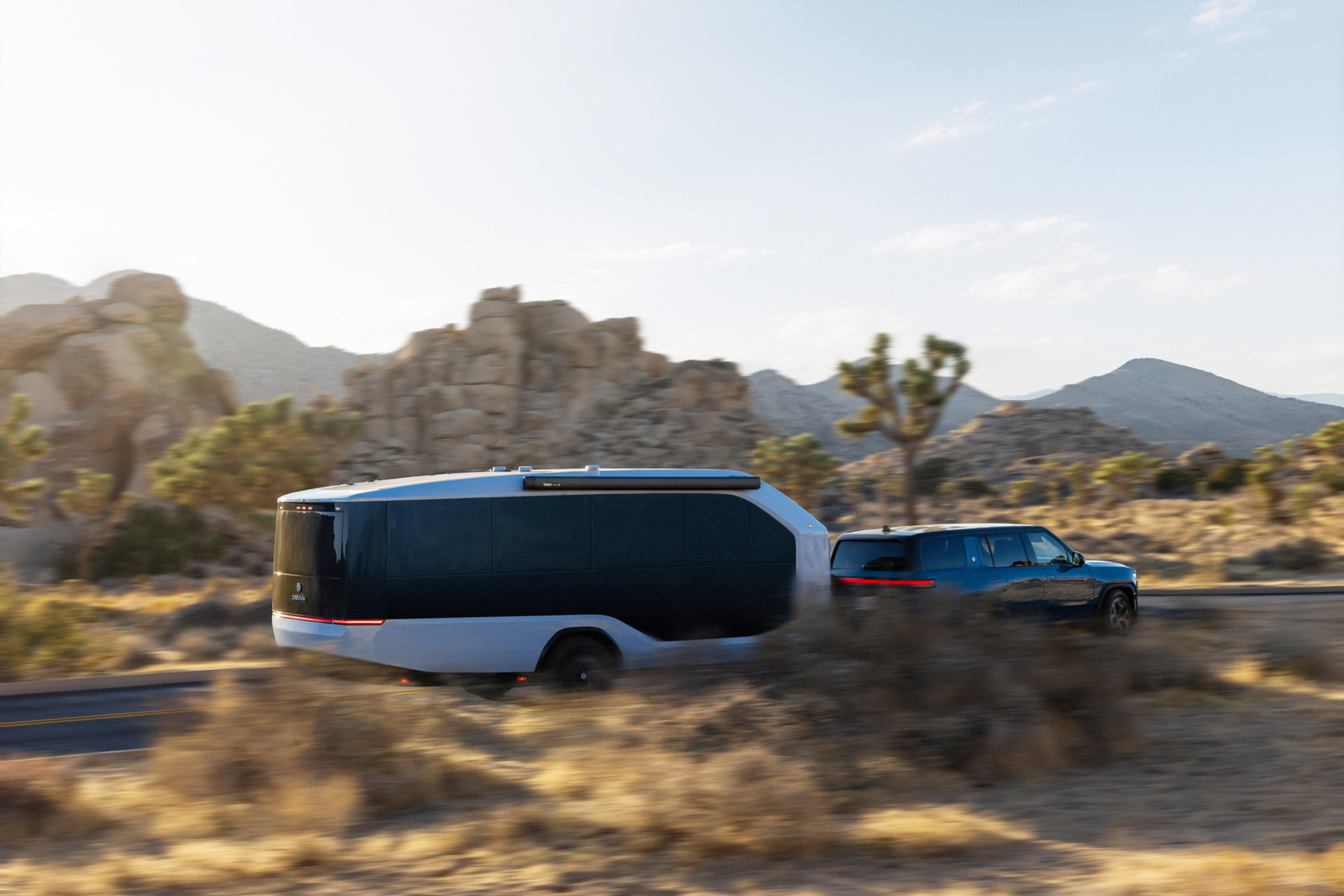A blue SUV is towing a sleek, modern trailer through a desert landscape with rocky hills and sparse vegetation.