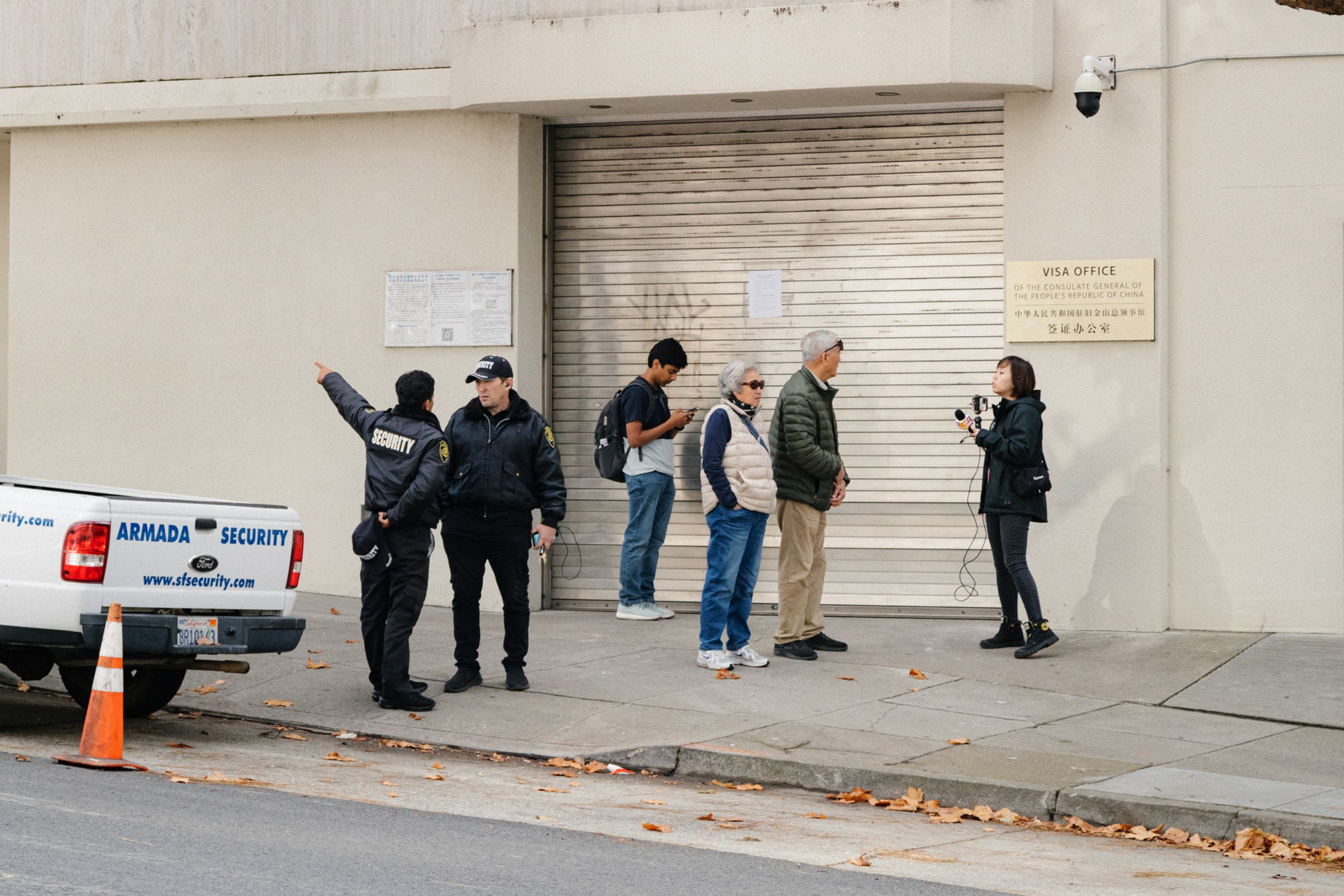 Two security guards stand by a truck while four people wait near a closed metal shutter at a visa office, one person using a phone.